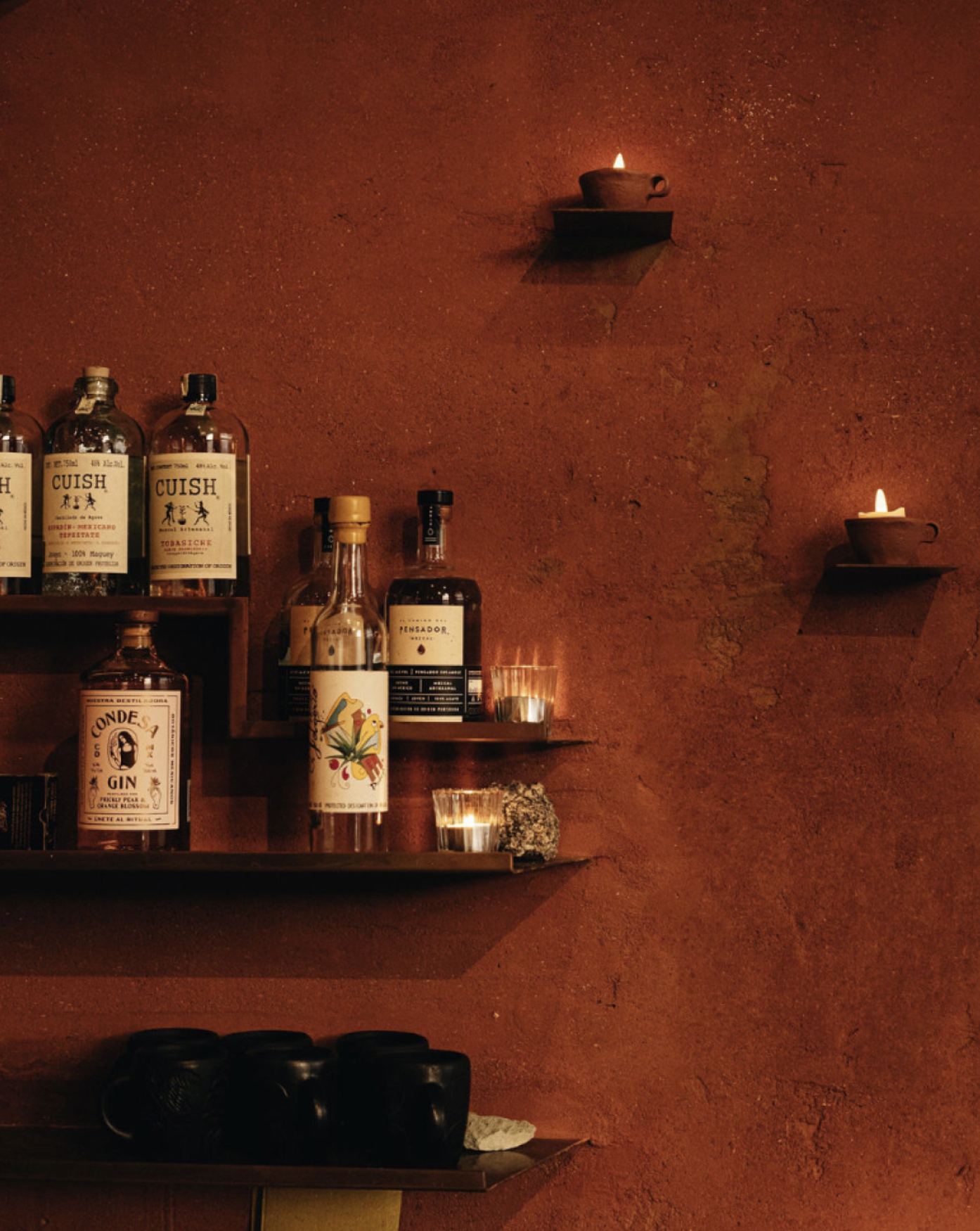 A wall with two small lit candles in clay holders and a shelf with various bottles of alcohol, including gin, tequila, and whiskey, against a textured reddish-brown wall.