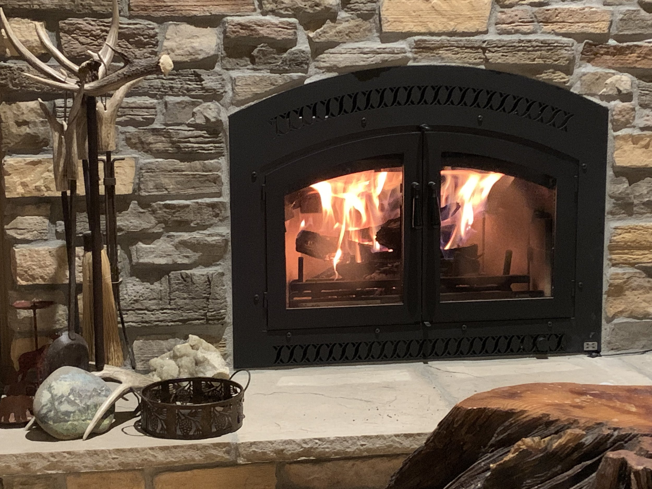 Fire burning inside a black metal fireplace with a glass door, set in a stone wall. To the left of the fireplace, there are fireplace tools, decorative stones, and a woven basket on a stone mantel.