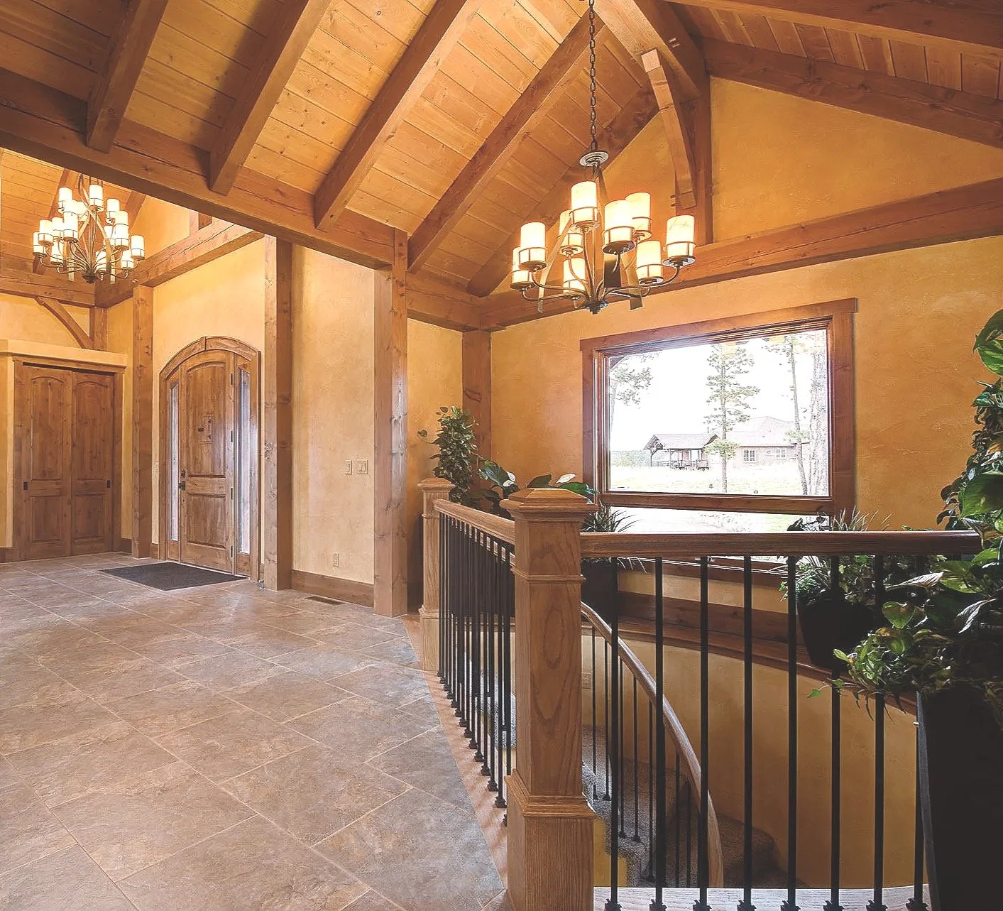 Interior view of a house hallway with stone tile flooring, wooden beams on the ceiling, a staircase with a wooden and black metal railing, and a large window showing trees and a house outside. There are chandeliers hanging from the ceiling and potted plants near the window.