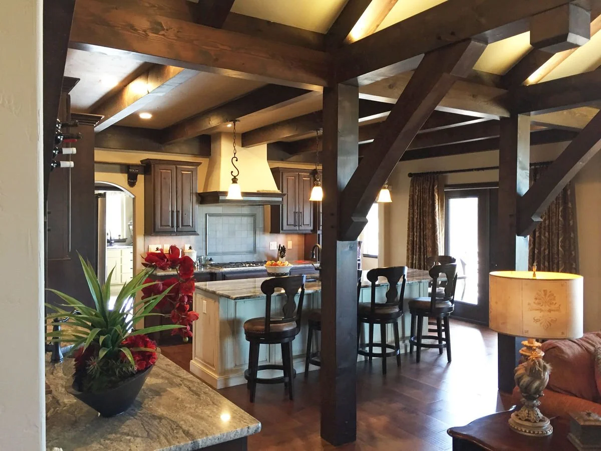Interior of a kitchen with dark wooden beams, a kitchen island with bar stools, and a lit table lamp in a living room area.