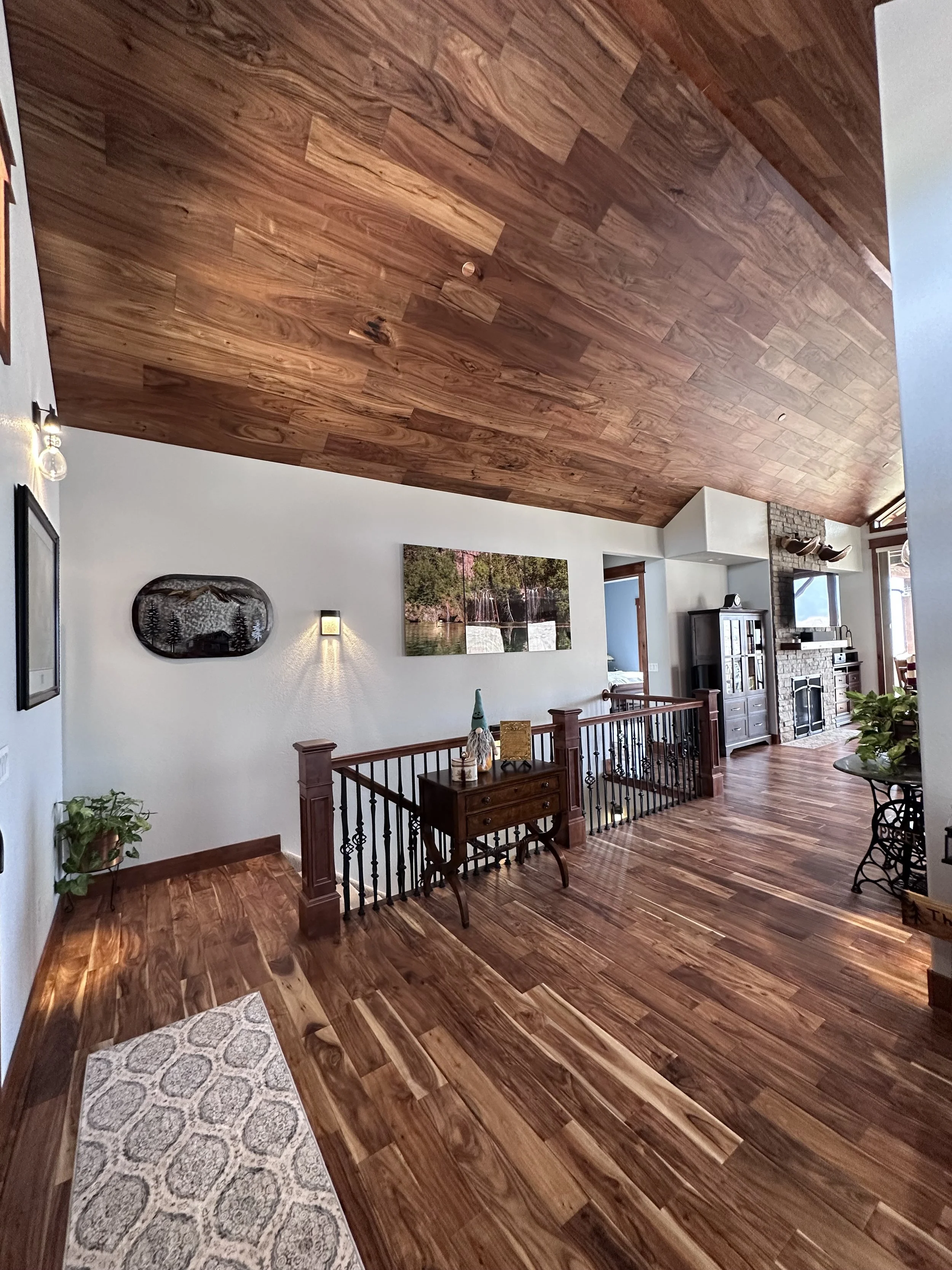 Interior view of a house with wooden floors and ceiling, white walls, and a staircase with a wooden and metal railing leading down. There is a small table with decorative items, a wall-mounted light, a large landscape photo, a cabinet, a fireplace, mounted TV, and a corner plant.
