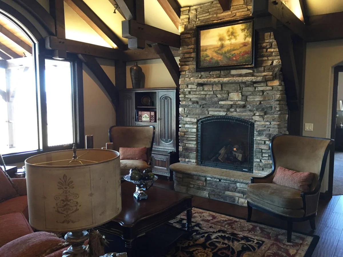 Living room with a stone fireplace, framed landscape painting above fireplace, two armchairs, a coffee table with decorative objects, a table lamp, and large windows letting in natural light.