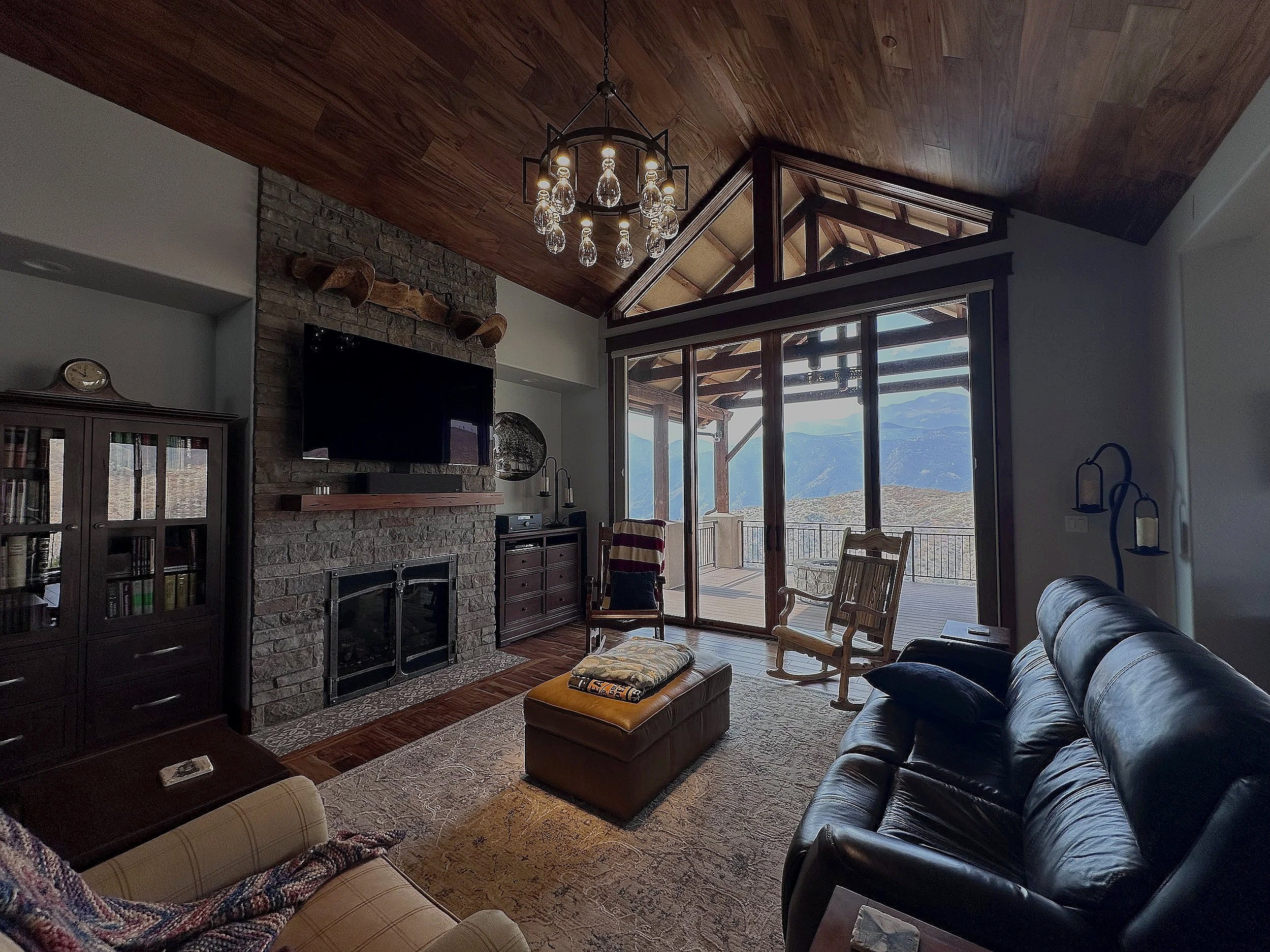 Living room with a stone fireplace, wooden ceiling, and large glass sliding doors. Outdoor mountain view, furniture including a leather sofa, wooden rocking chairs, and a cushioned ottoman.