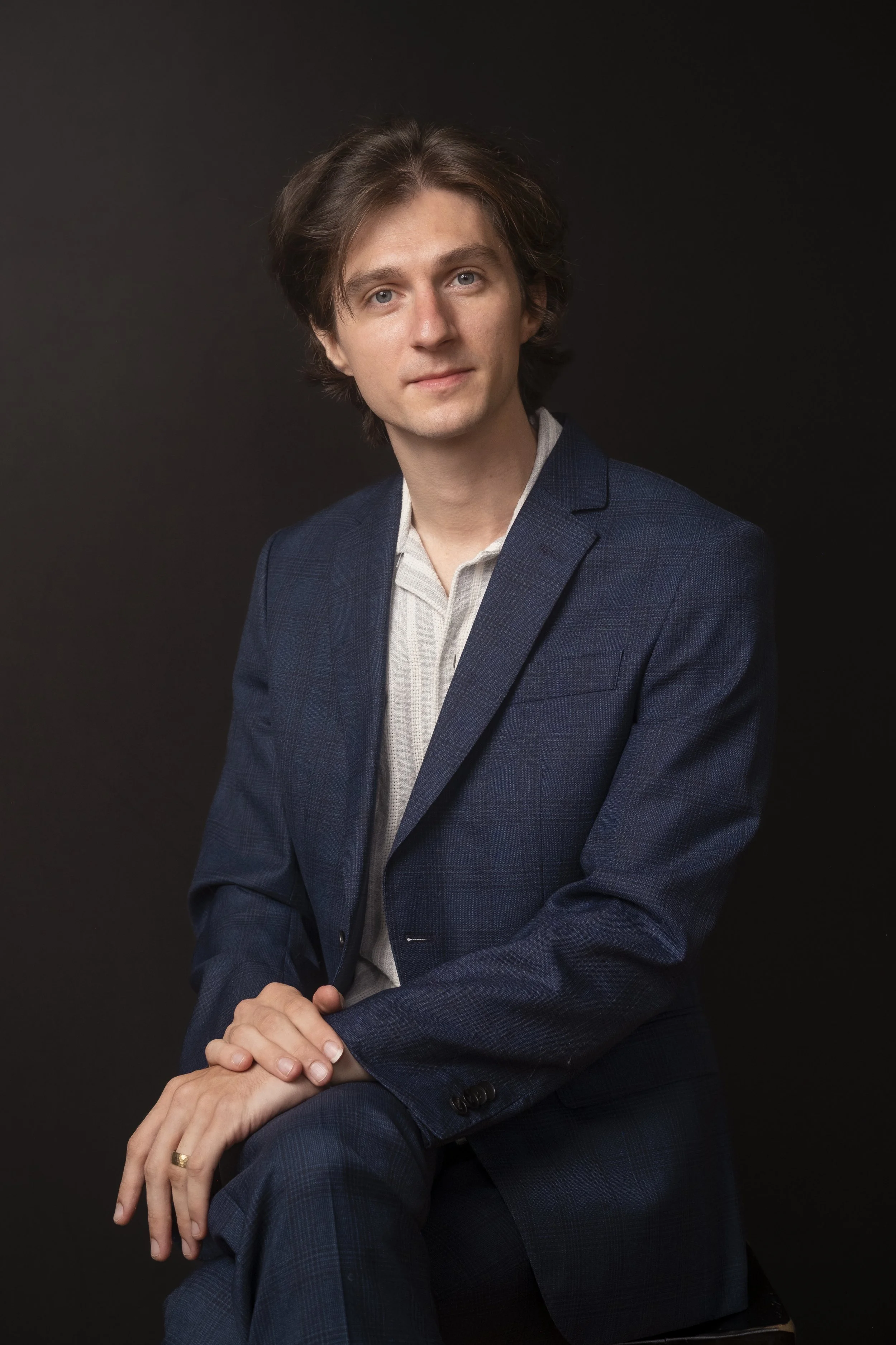 Portrait of Trevin Nelson, multi-instrumentalist and educator with wavy brown hair, blue eyes, and fair skin, dressed in a dark blue suit with a light gray shirt, seated against a black background, looking directly at the camera.