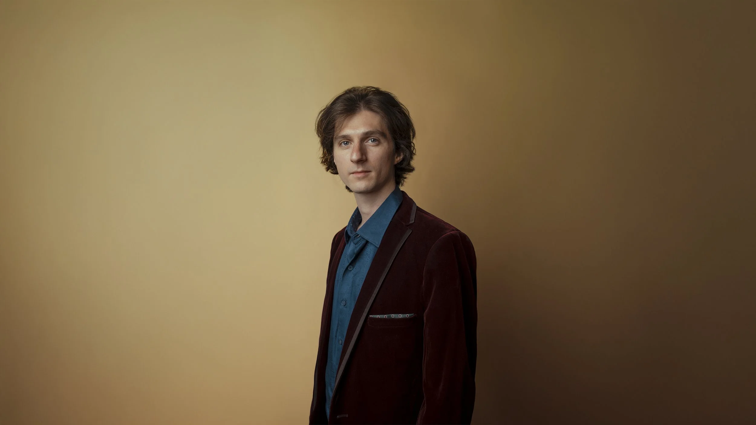 Portrait of Trevin Nelson, banjoist and multi-instrumentalist with medium-length brown hair, wearing a dark red blazer and blue shirt, standing against a gradient gold background.