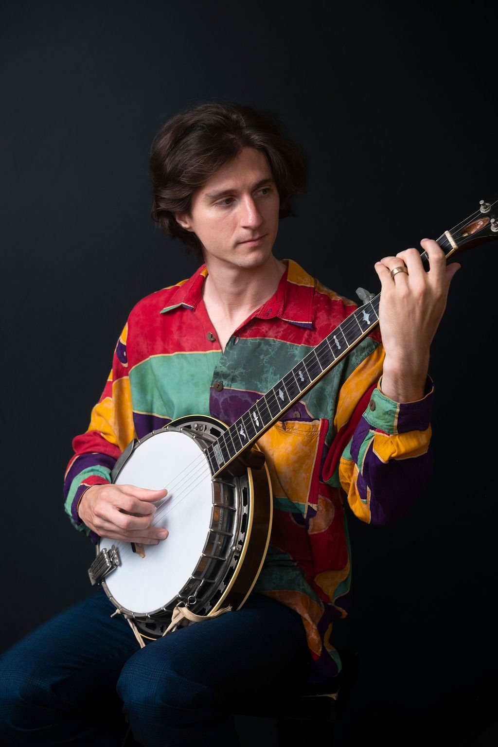 A young man with dark, voluminous hair playing a banjo against a plain dark background.