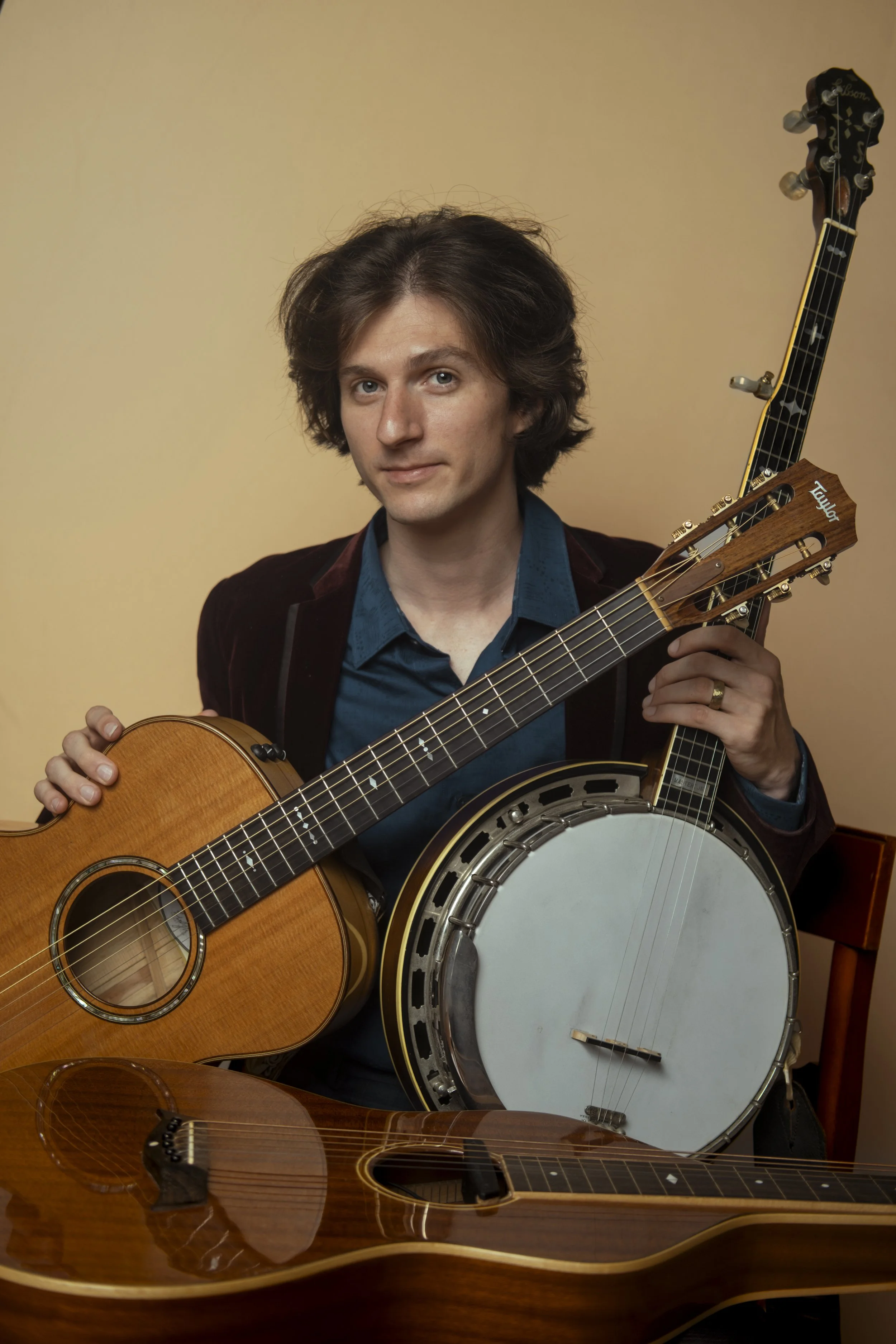 Portrait of Trevin Nelson, multi-instrumentalist and educator with medium-length dark hair, wearing a dark red jacket and blue shirt, holding a banjo, acoustic guitar, and lap steel slide guitar sitting against a plain gold background.