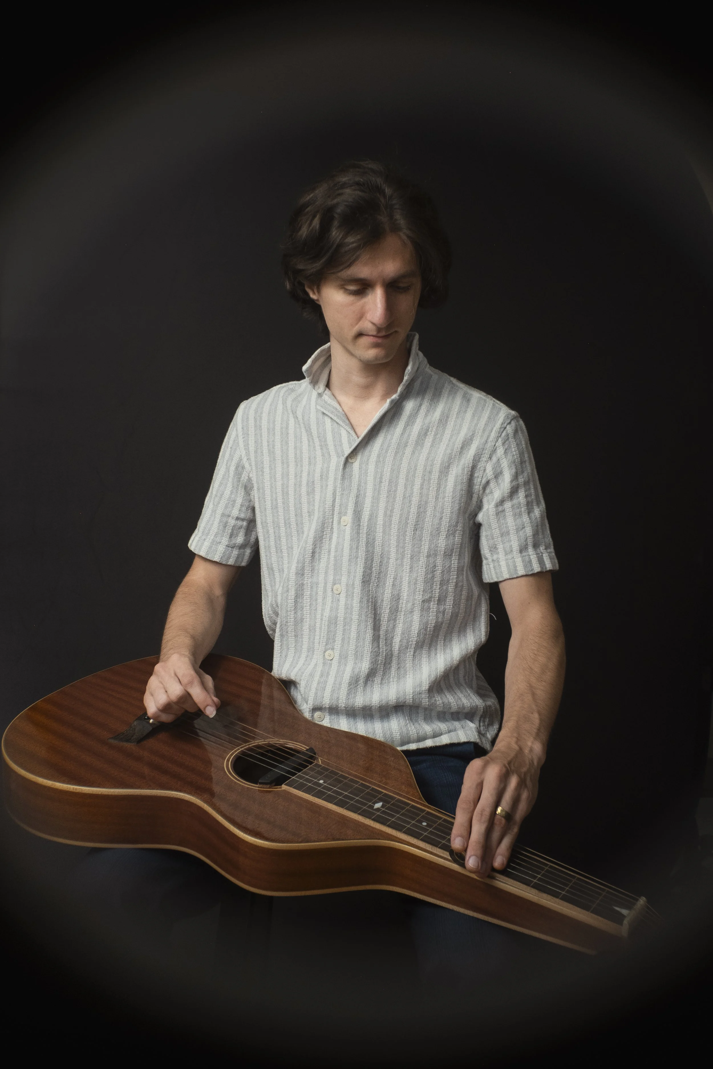 Portrait of Trevin Nelson, multi-instrumentalist and educator with dark, wavy hair wearing a light gray striped button-up shirt playing a lap steel slide guitar against a dark background.