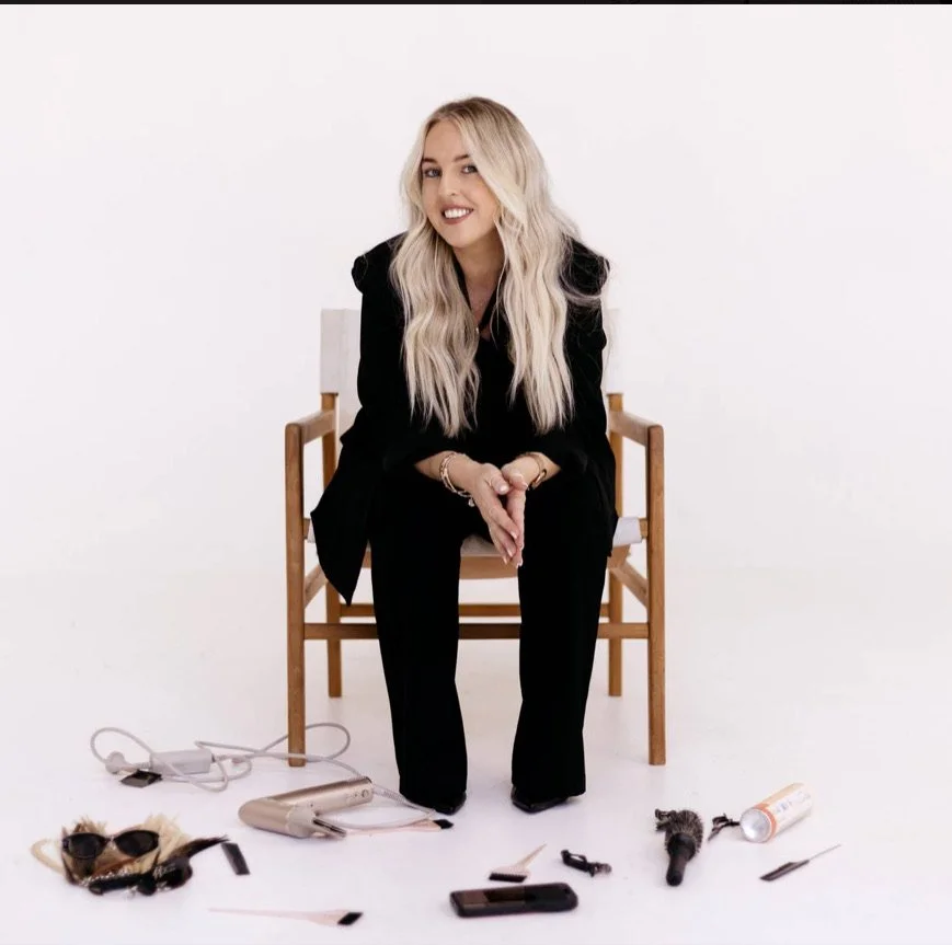 Woman sitting on chair with hair tools and styling accessories scattered on the floor in front of her, white background.
