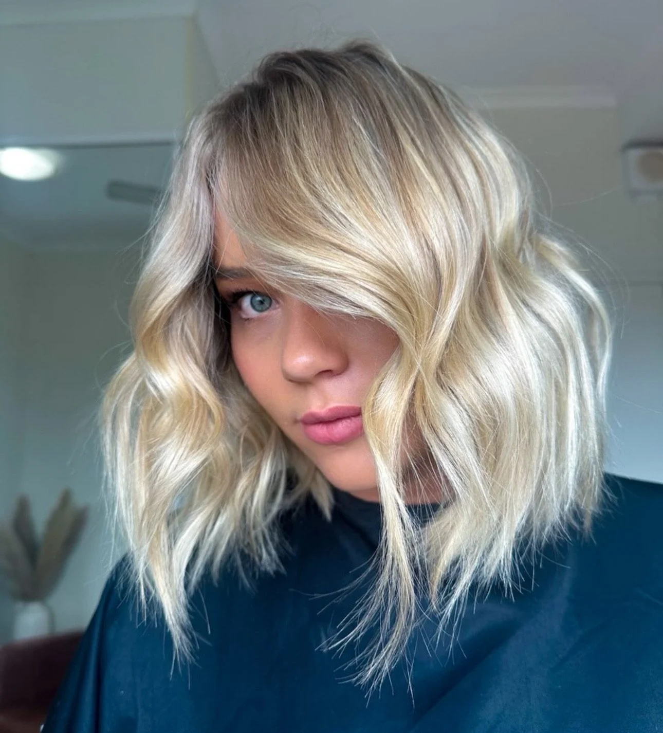 A woman with blonde, wavy hair partially covering her face, with striking blue eyes and wearing a black top, in a well-lit indoor setting.