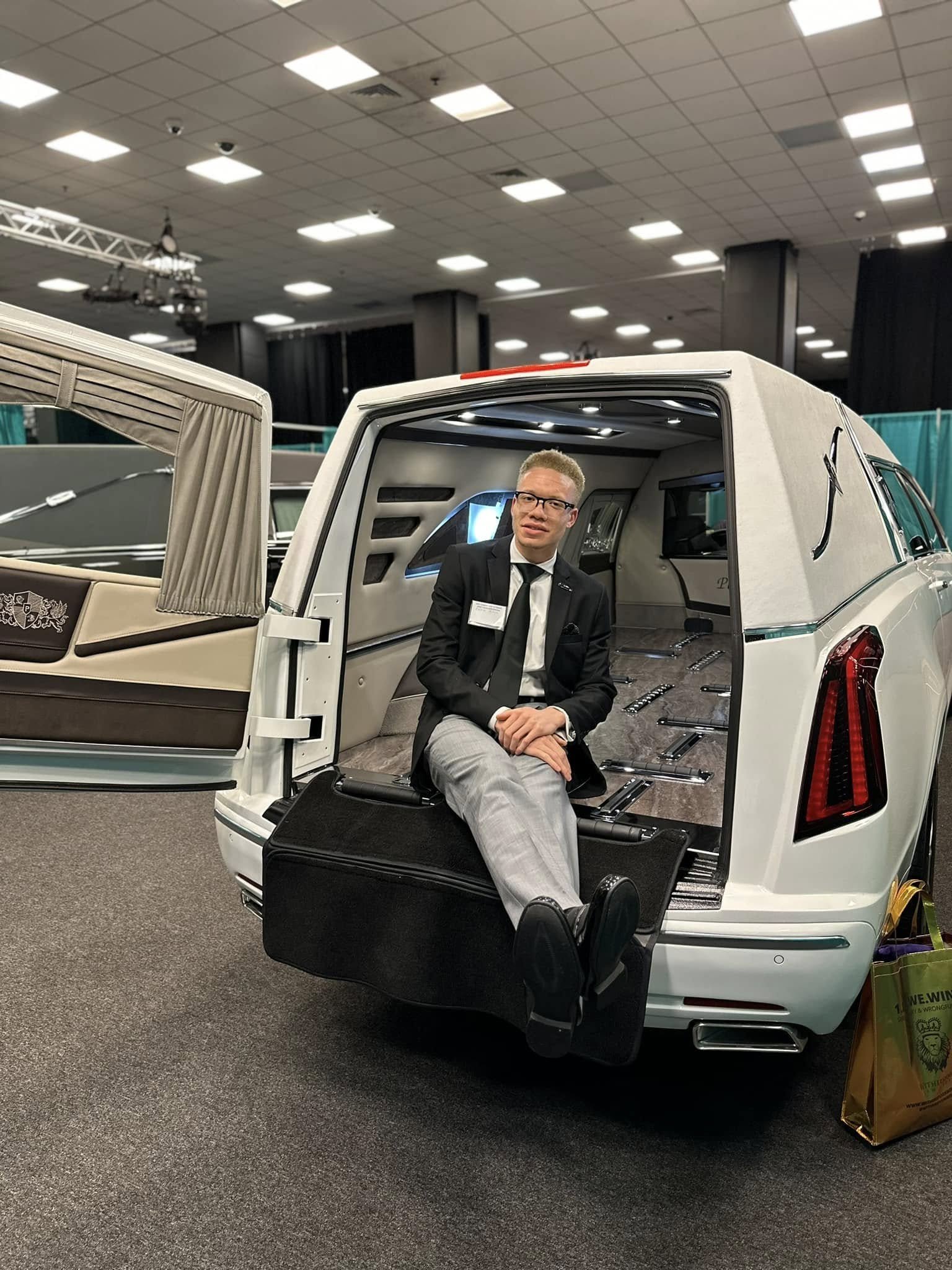 A man sitting on a black case in the open trunk of a white limousine at an indoor event or exhibition.