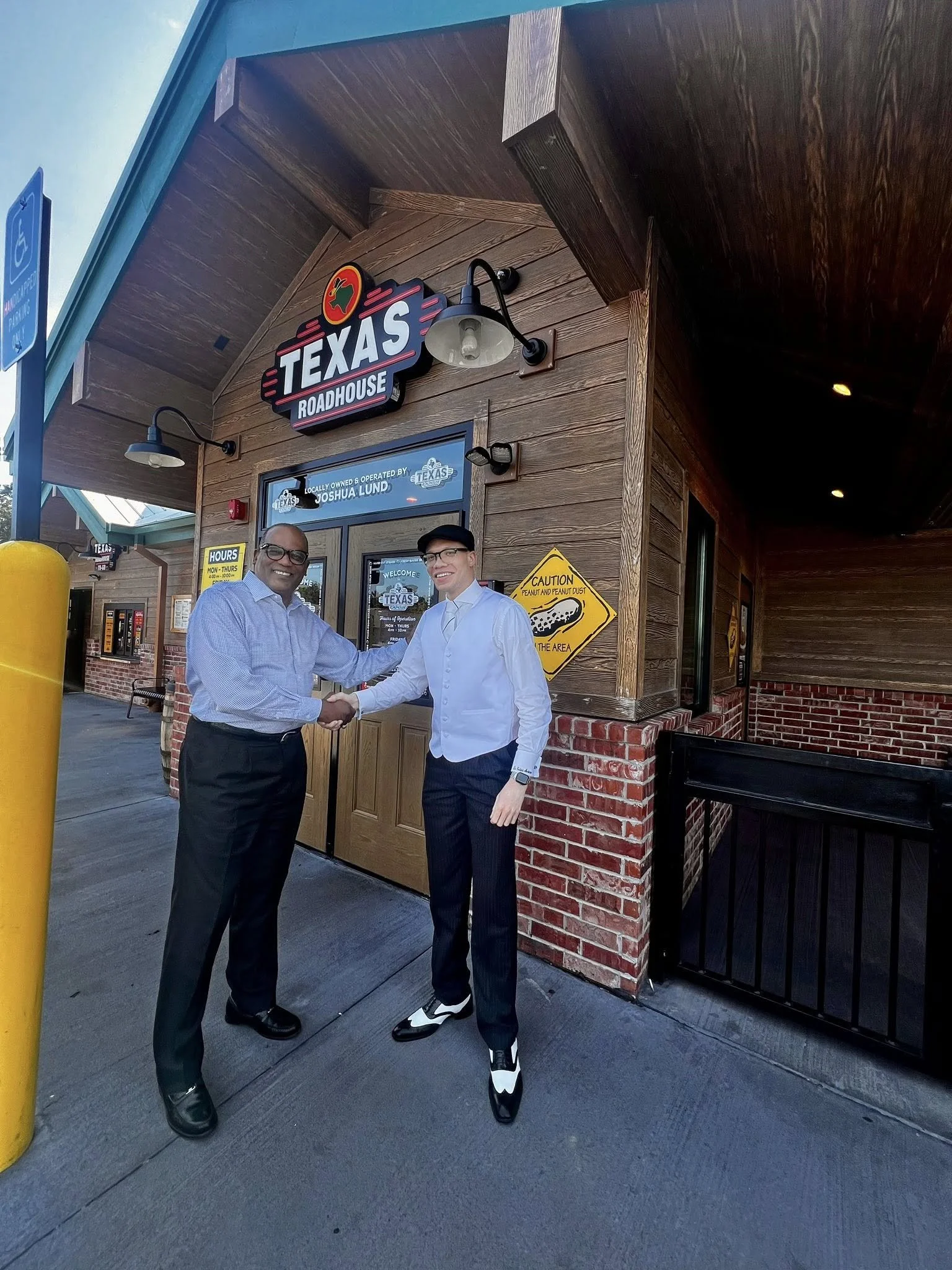 Two men shaking hands outside Texas Roadhouse restaurant. One is wearing glasses and a blue shirt, the other in a white shirt, black cap, and dress shoes. The restaurant sign is visible above and the building has a wood and brick exterior.