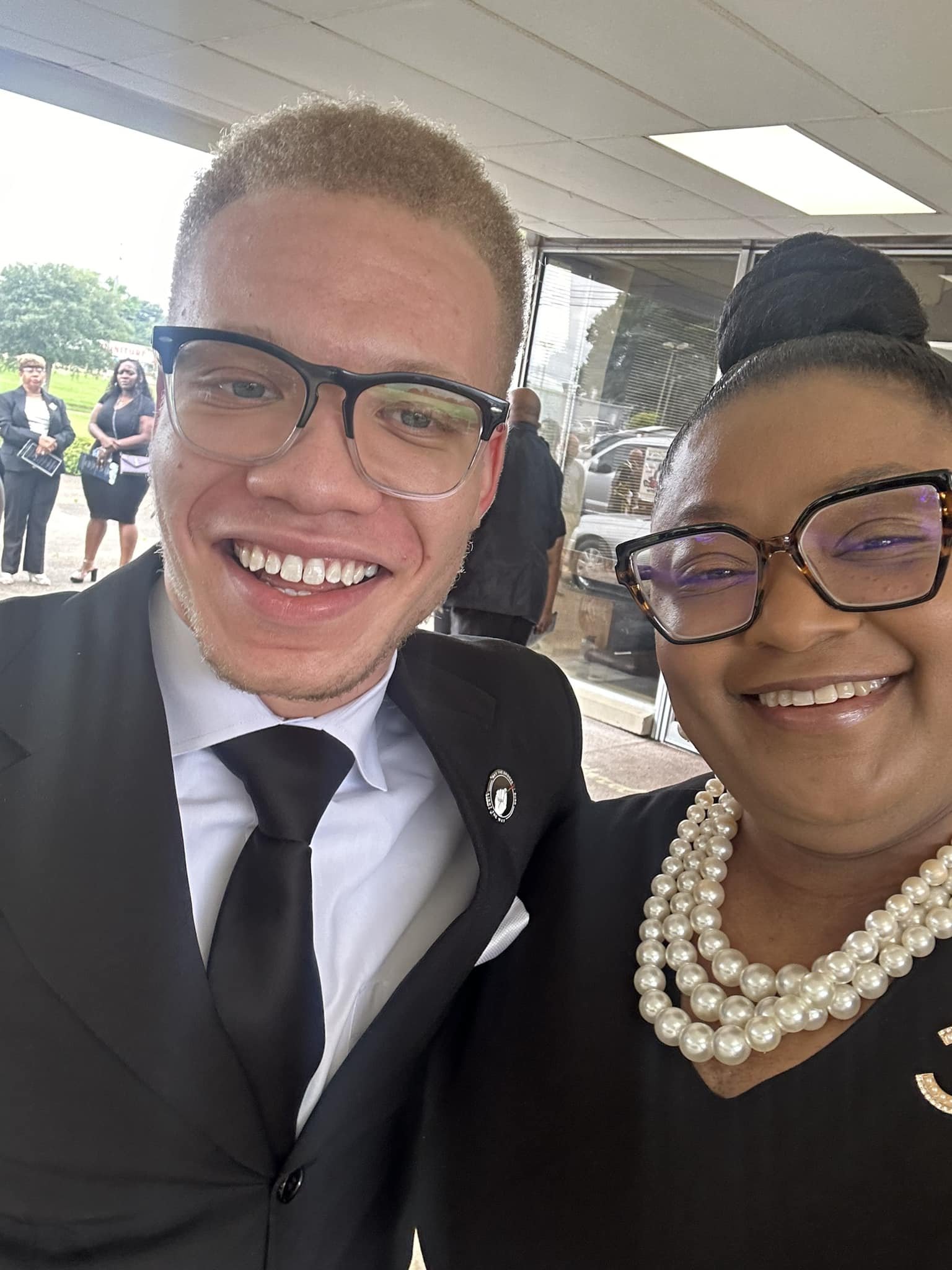 A smiling young man in a black suit, white shirt, and black tie taking a selfie with a woman wearing glasses, a pearl necklace, and a black dress in front of a building with glass windows and open doors.