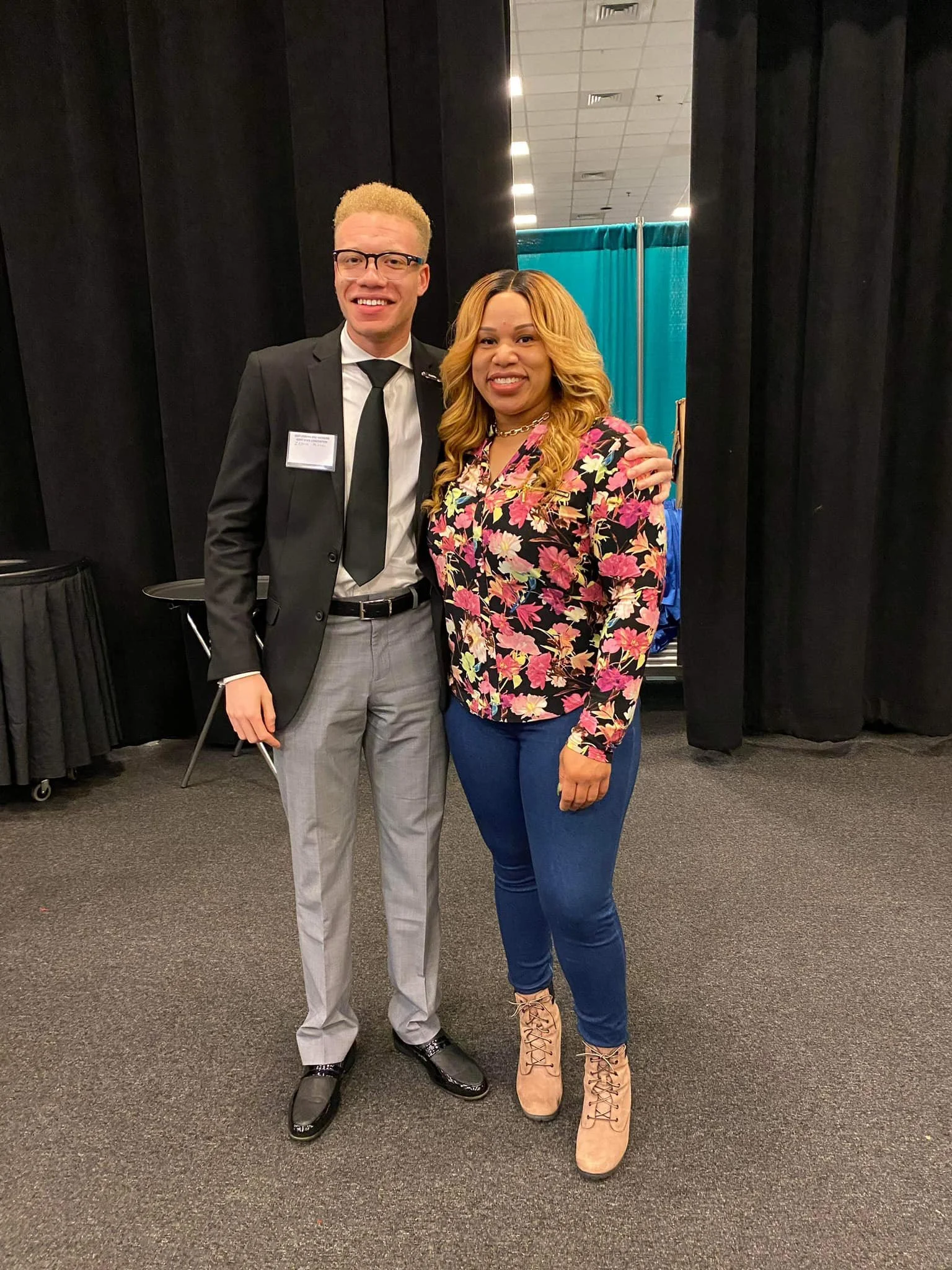 A man and woman standing together in a conference or event setting, smiling at the camera. The man is wearing a black blazer, white shirt, black tie, gray pants, and black shiny shoes. The woman is wearing a colorful floral blouse, blue jeans, and be