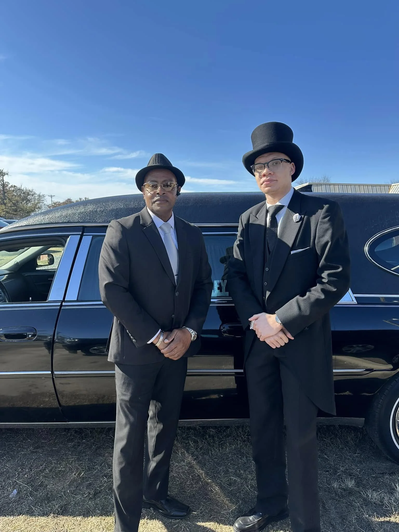 Two men dressed in black suits, ties, and hats standing in front of a black limo car on a sunny day.