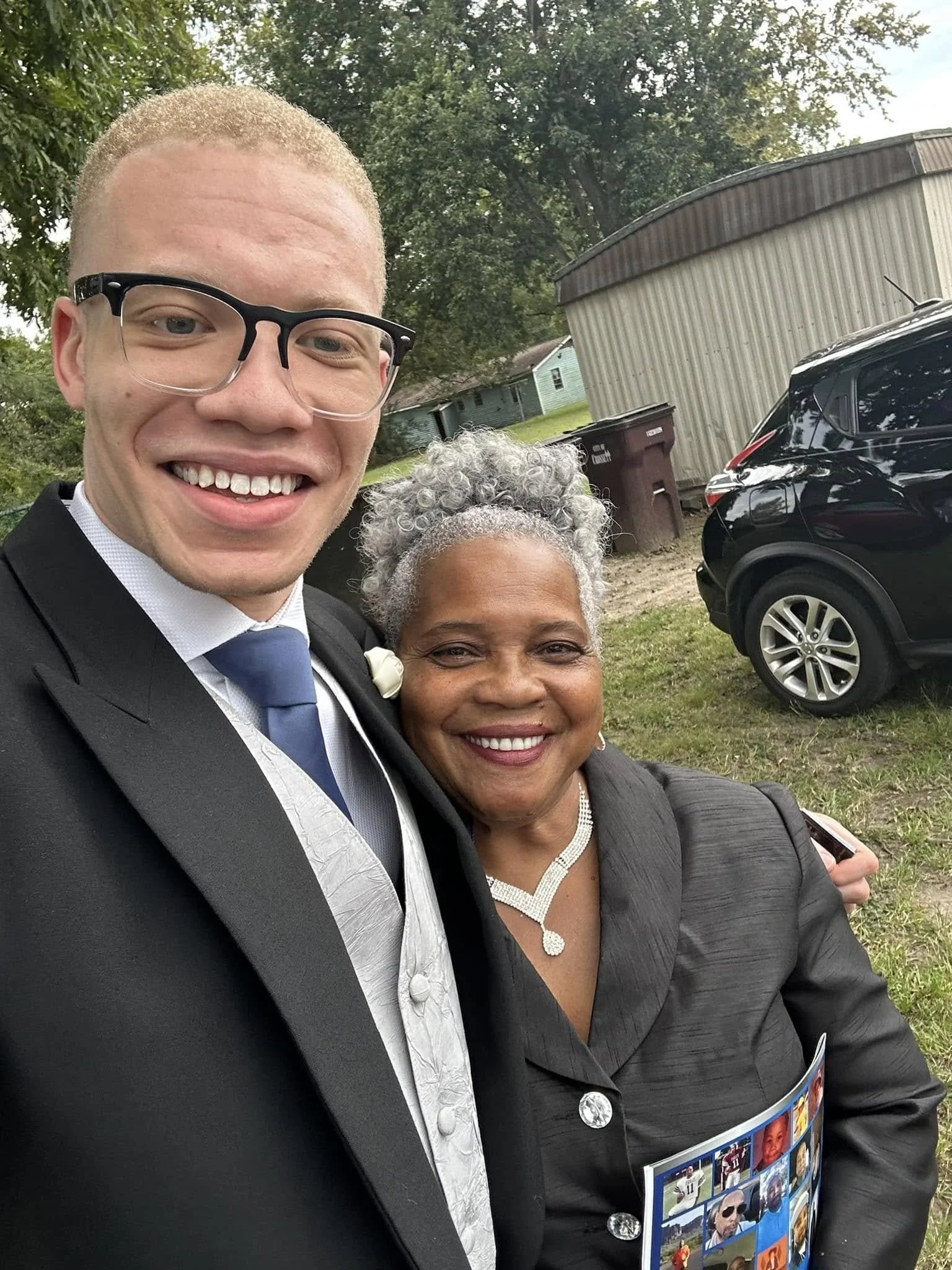 A young man and an older woman smiling and taking a selfie outdoors. The young man is wearing a black suit, white shirt, and blue tie, and the older woman is dressed in a black outfit with a white necklace and has gray curly hair. There are trees, a 