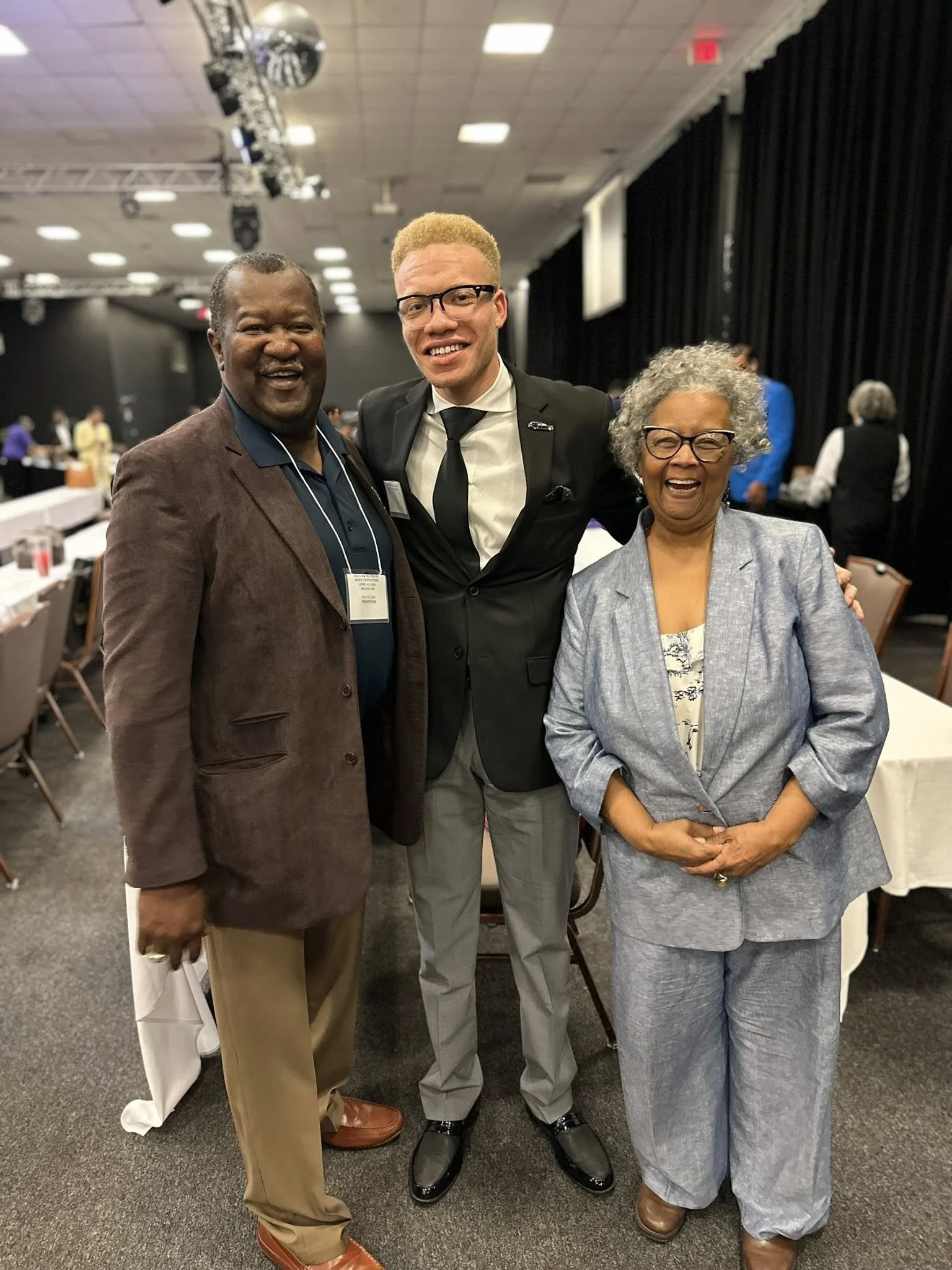 Three people standing together at an event, smiling for the photo. The person in the center is wearing a black suit and tie, and the others are dressed in business casual attire. The background shows a conference or banquet hall with tables and other