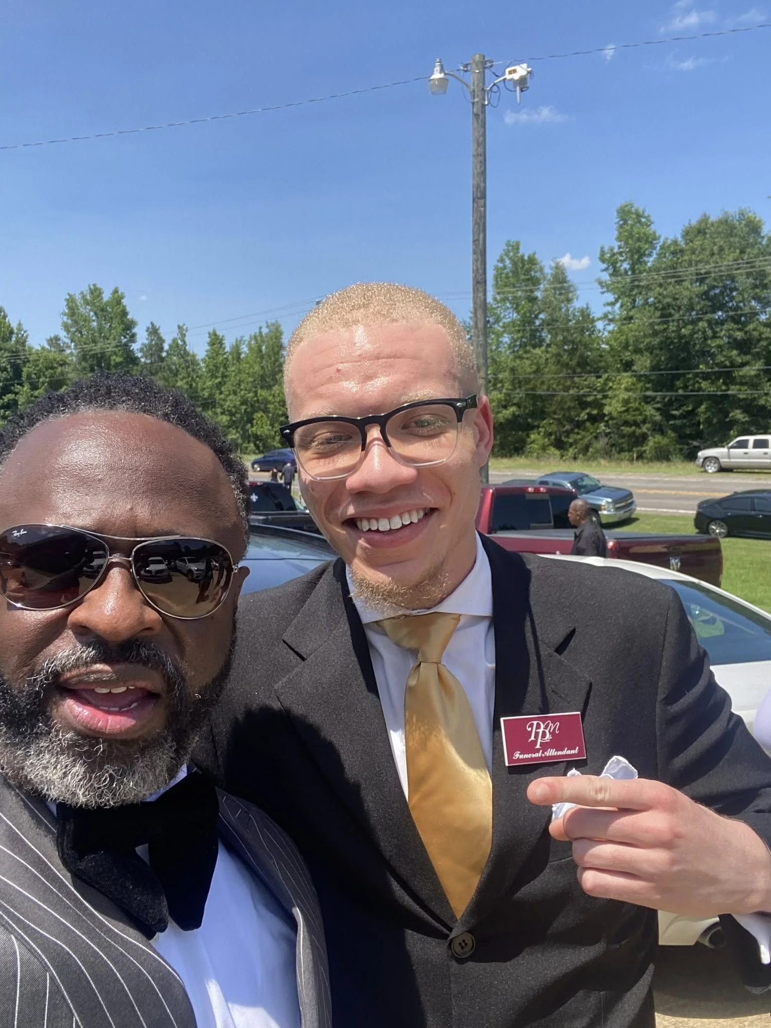 Two men dressed in formal attire taking a selfie outdoors on a sunny day, with cars and a power line in the background.