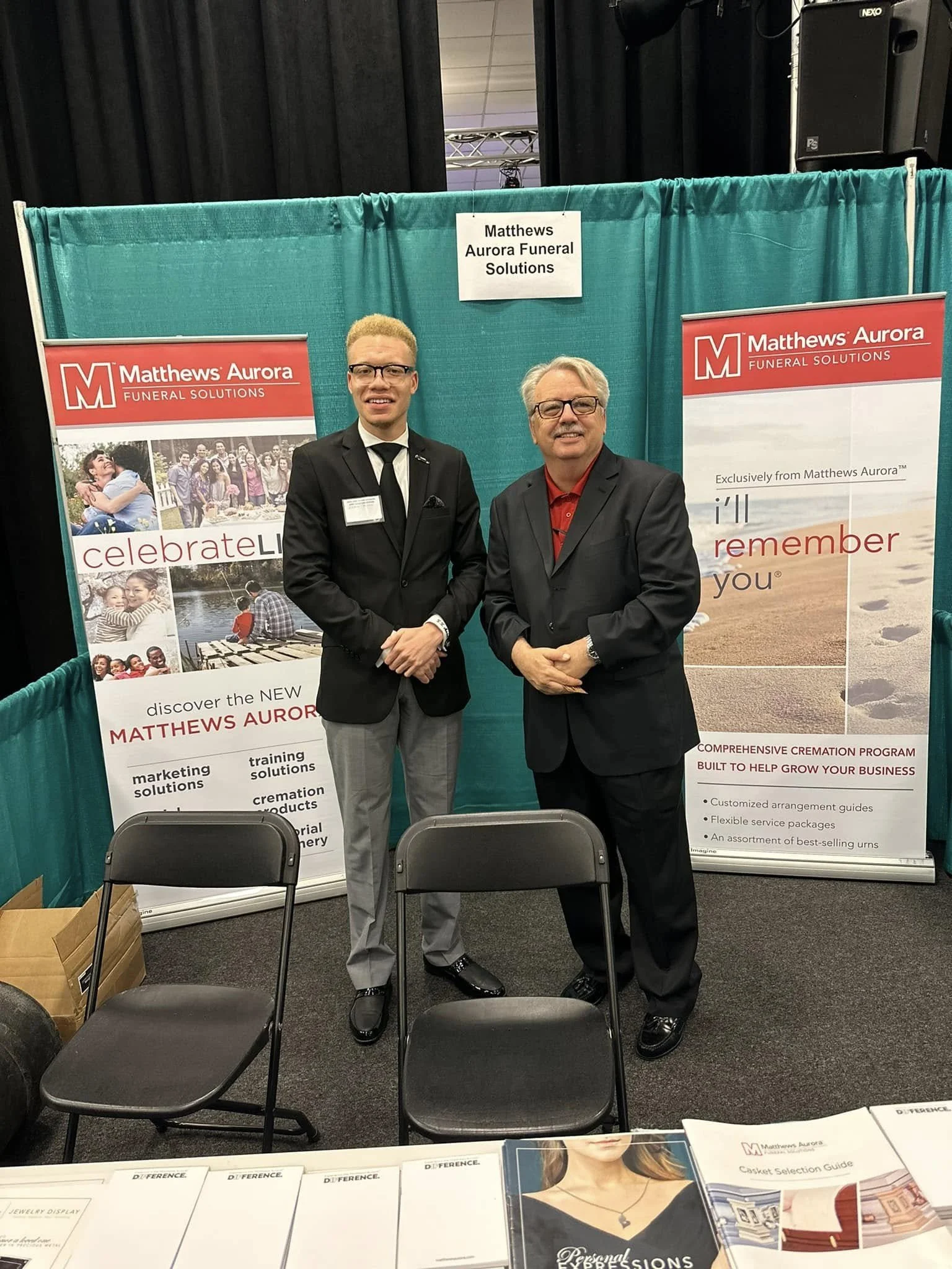 Two men standing in front of a booth for Matthews Aurora Funeral Solutions at an event. The booth has banners with the company's logo and services, and a table with pamphlets and brochures.