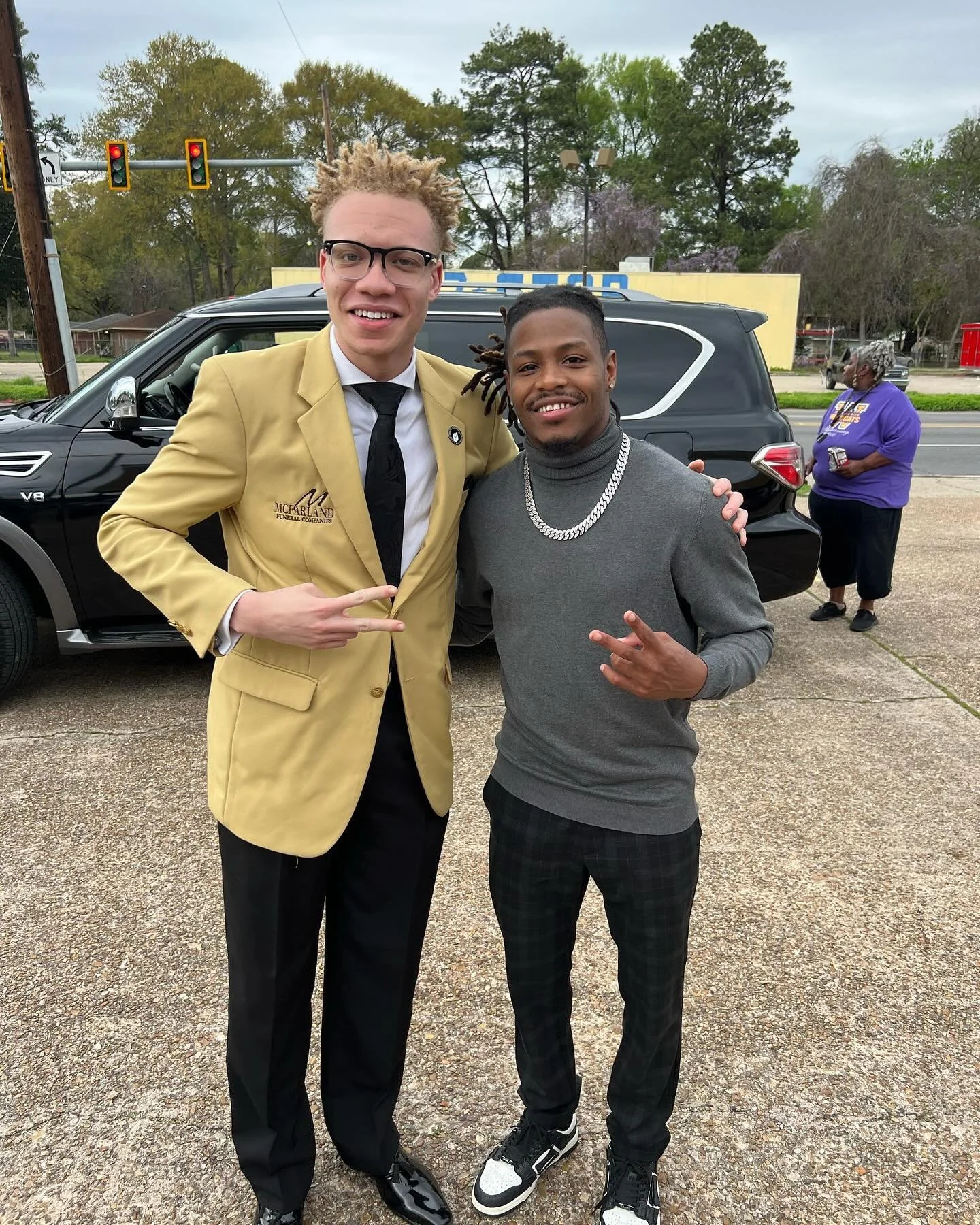 Two men posing for a photo outdoors, one wearing a yellow suit jacket and black tie, and the other in a gray turtleneck and chain necklace. They are smiling and making hand gestures. In the background, a woman in purple holds a drink near a black SUV