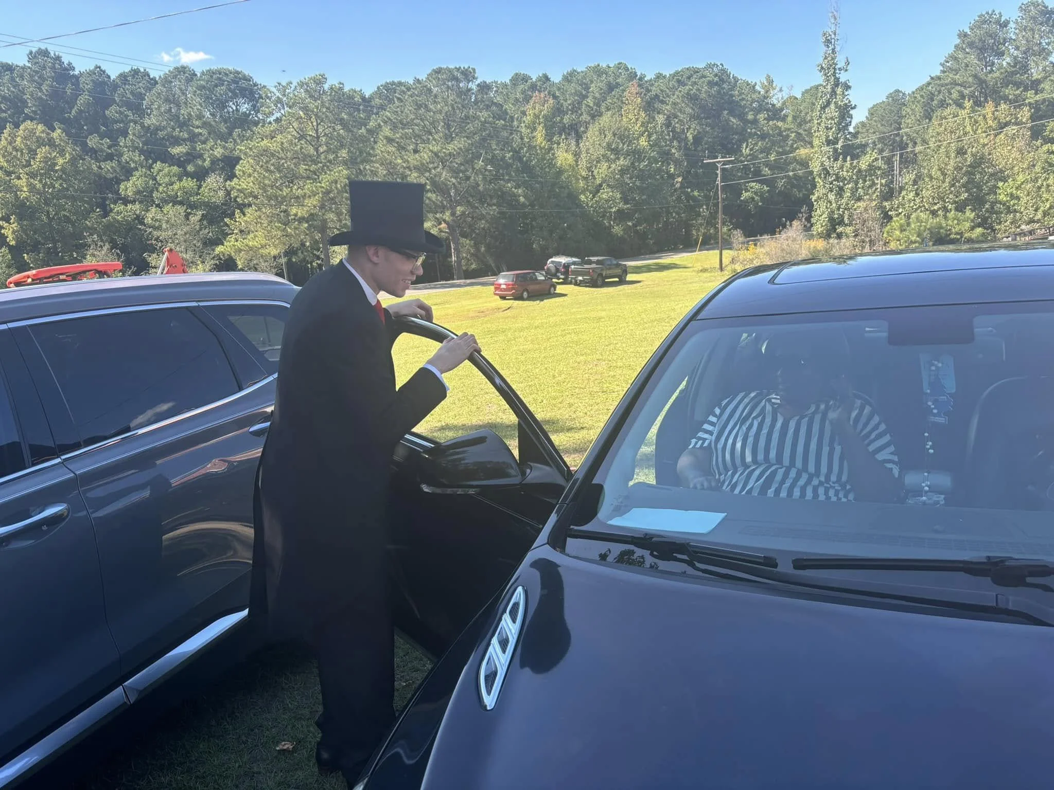 A man dressed as Abraham Lincoln, wearing a black suit, tie, and tall black hat, standing outside and talking to a woman inside a black car. The woman is wearing a black and white striped shirt and sunglasses, and is sitting in the driver's seat. The