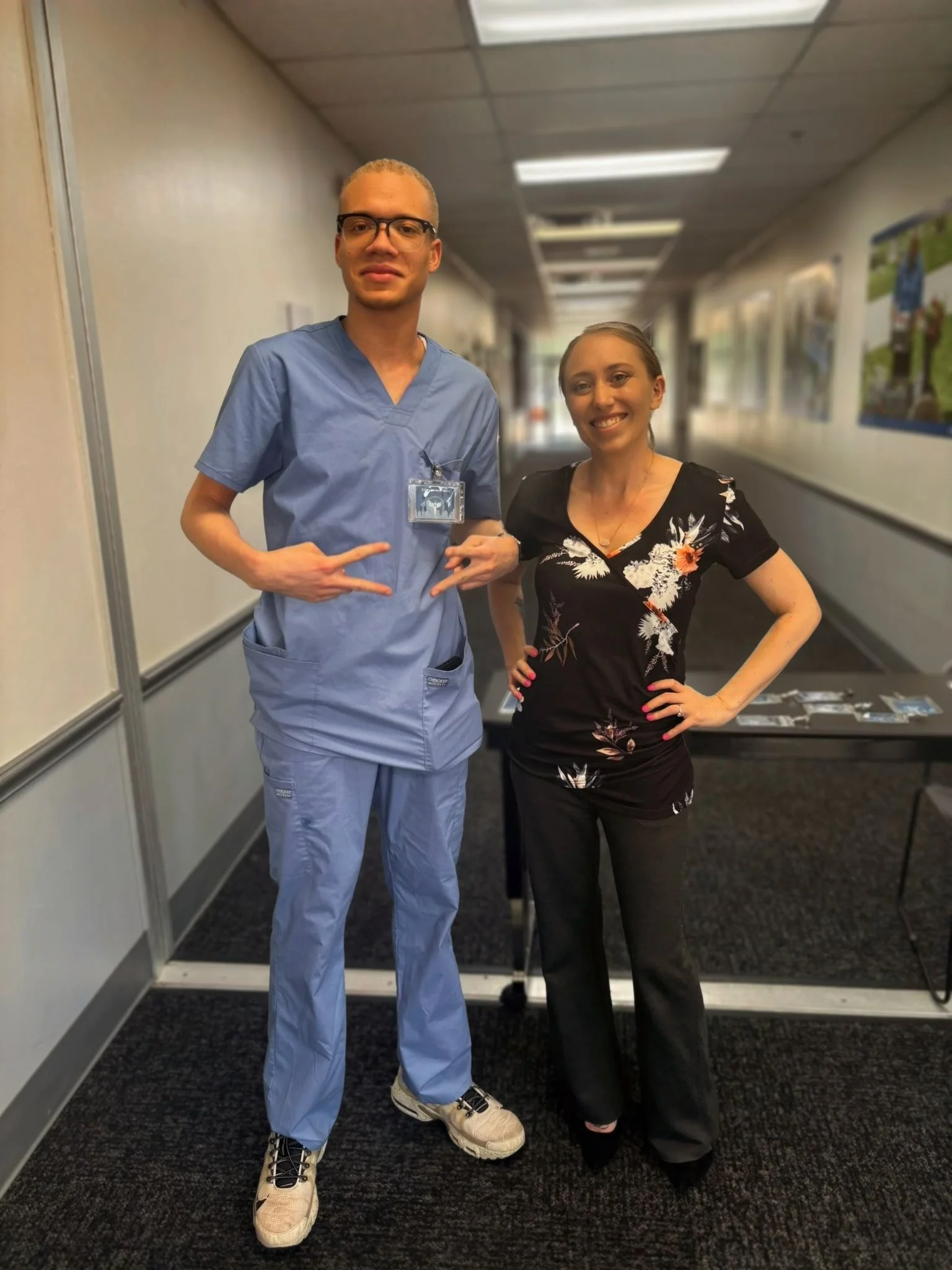 A young man in medical scrubs standing next to a woman in a floral blouse in a hallway.