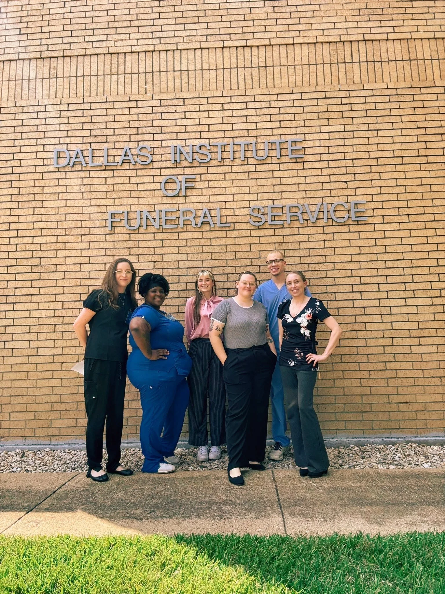 Group of six diverse women standing in front of a brick wall with a sign that reads 'Dallas Institute of Funeral Service'.