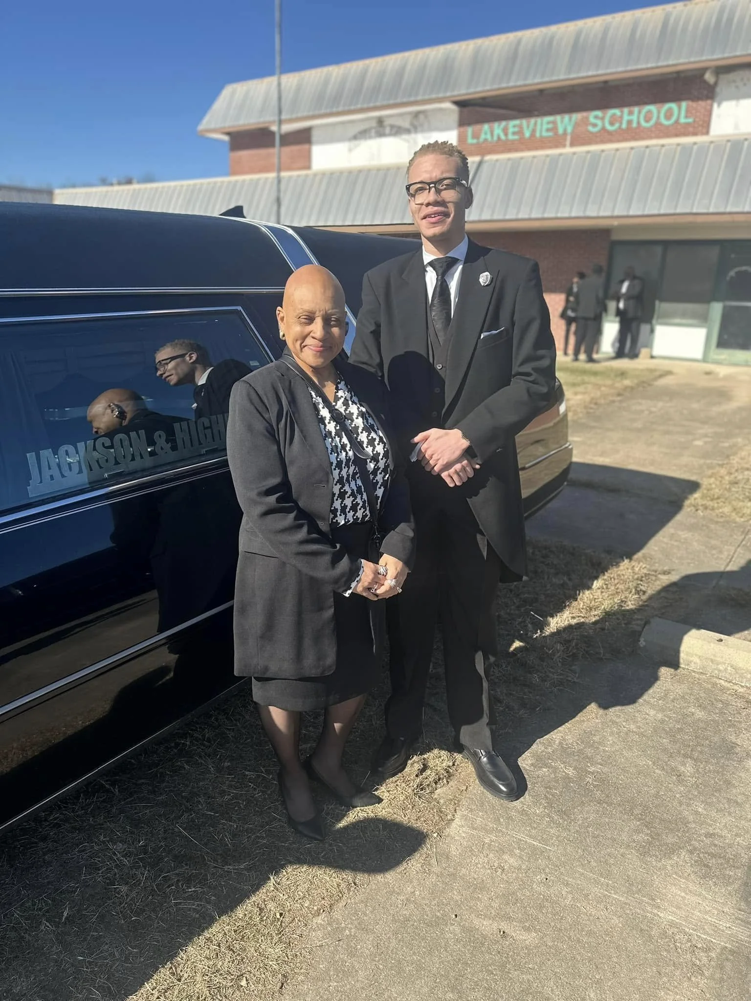 A woman and a man dressed in formal attire standing outside on a sunny day in front of a black limousine with a sign that reads 'Jackson & High'. In the background, there is a building with a sign that says 'Lakeview School' and a few people dressed 