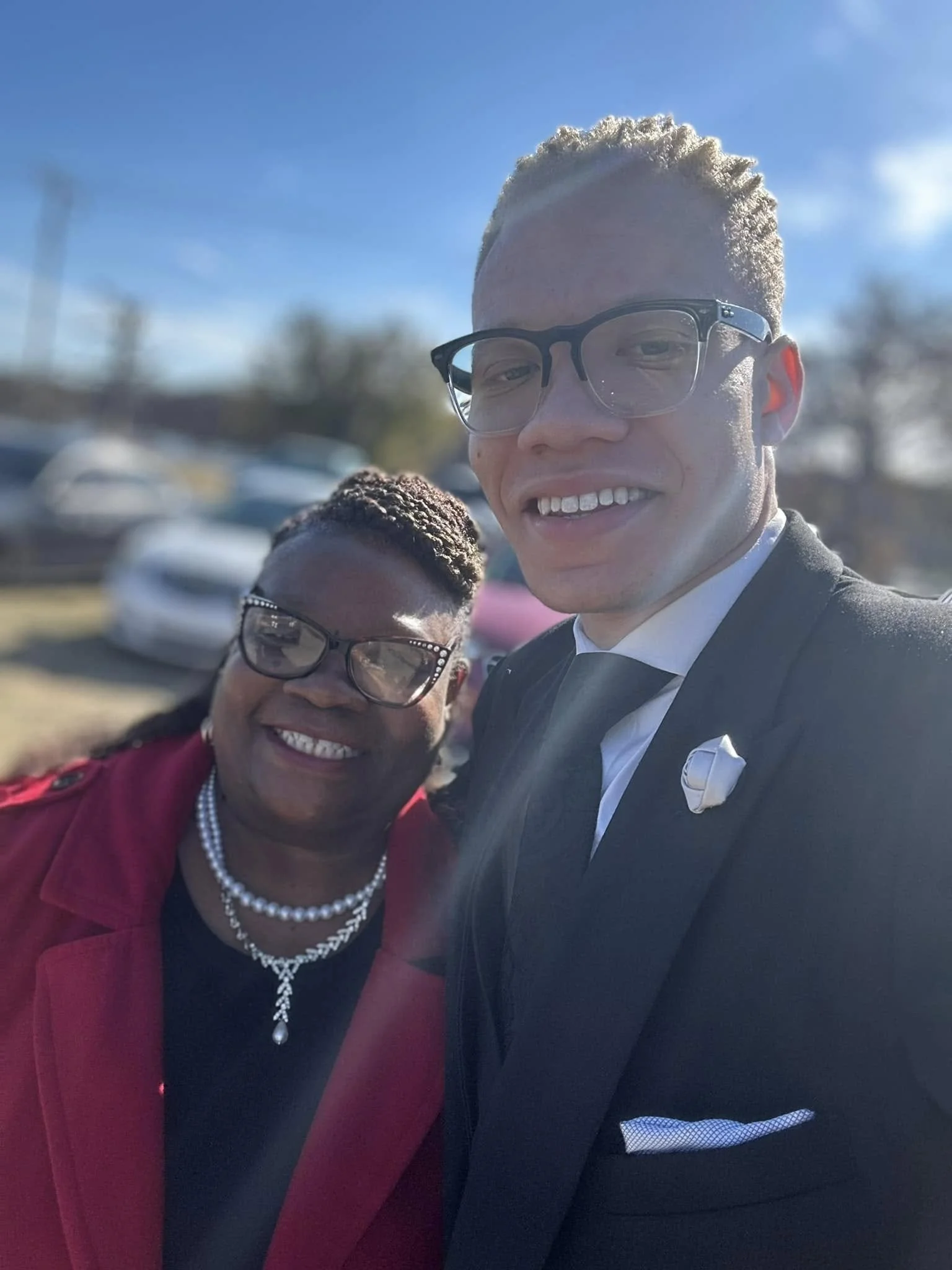 A smiling woman and a man are taking a selfie outdoors on a clear, sunny day. The woman is wearing glasses, a red coat, and a pearl necklace, while the man is dressed in a black suit with a white shirt and tie, with glasses, against a blurred backgro