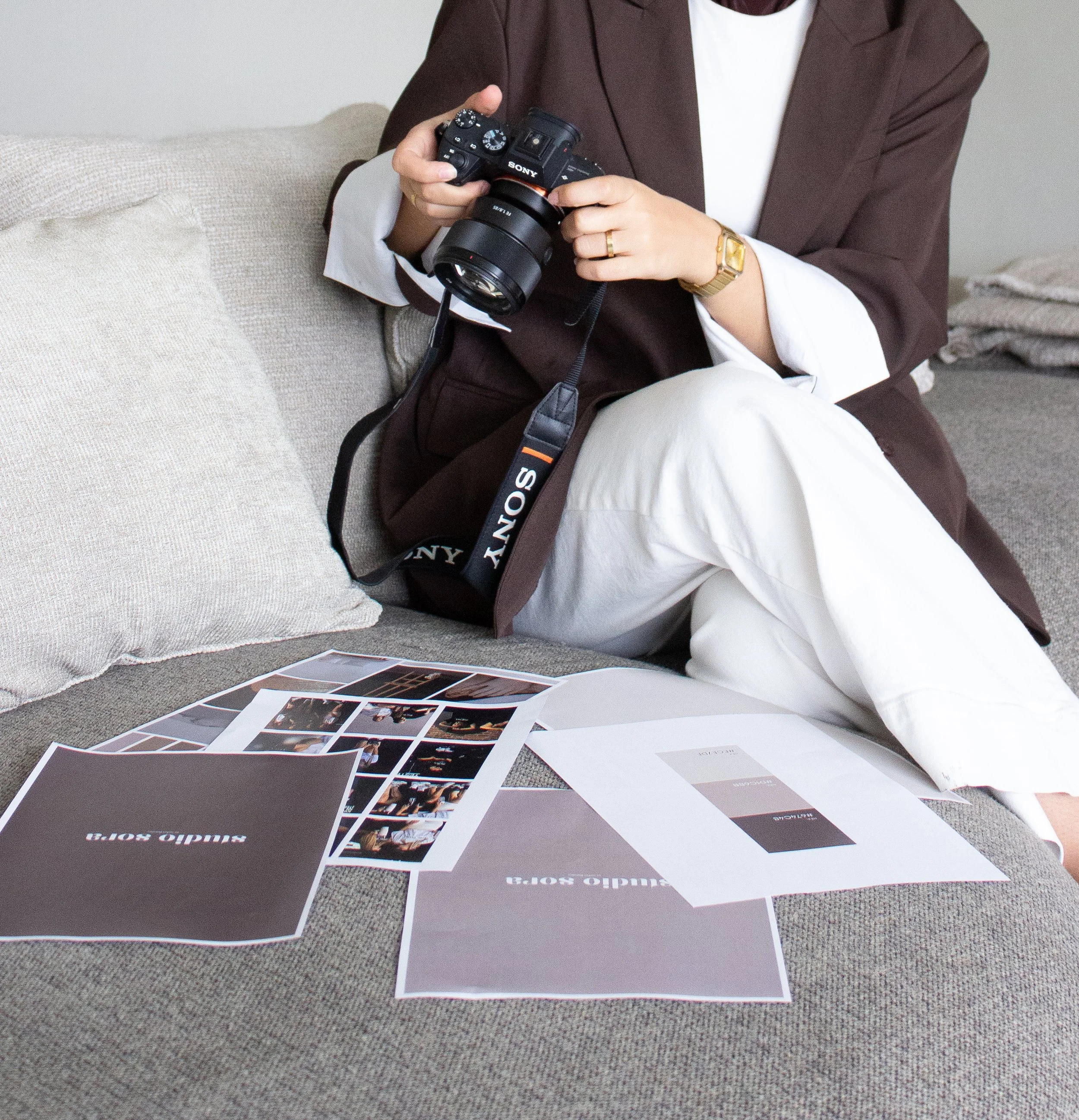 Person sitting on a beige sofa, holding a Sony camera, surrounded by printed photographs and color swatches related to a photo shoot or design project.