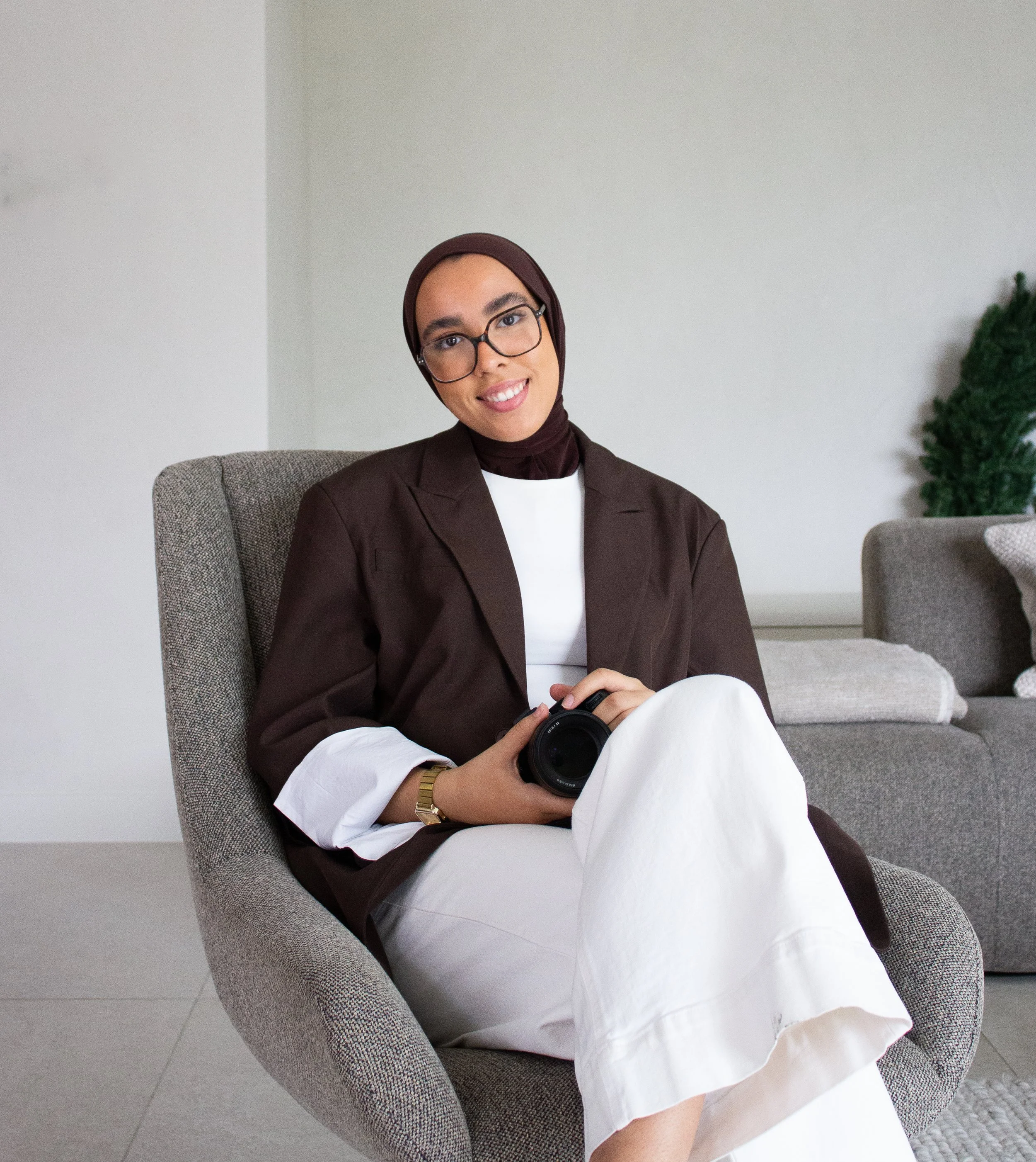 A woman with glasses and a hijab sitting in a modern living room, holding a camera, smiling at the camera.