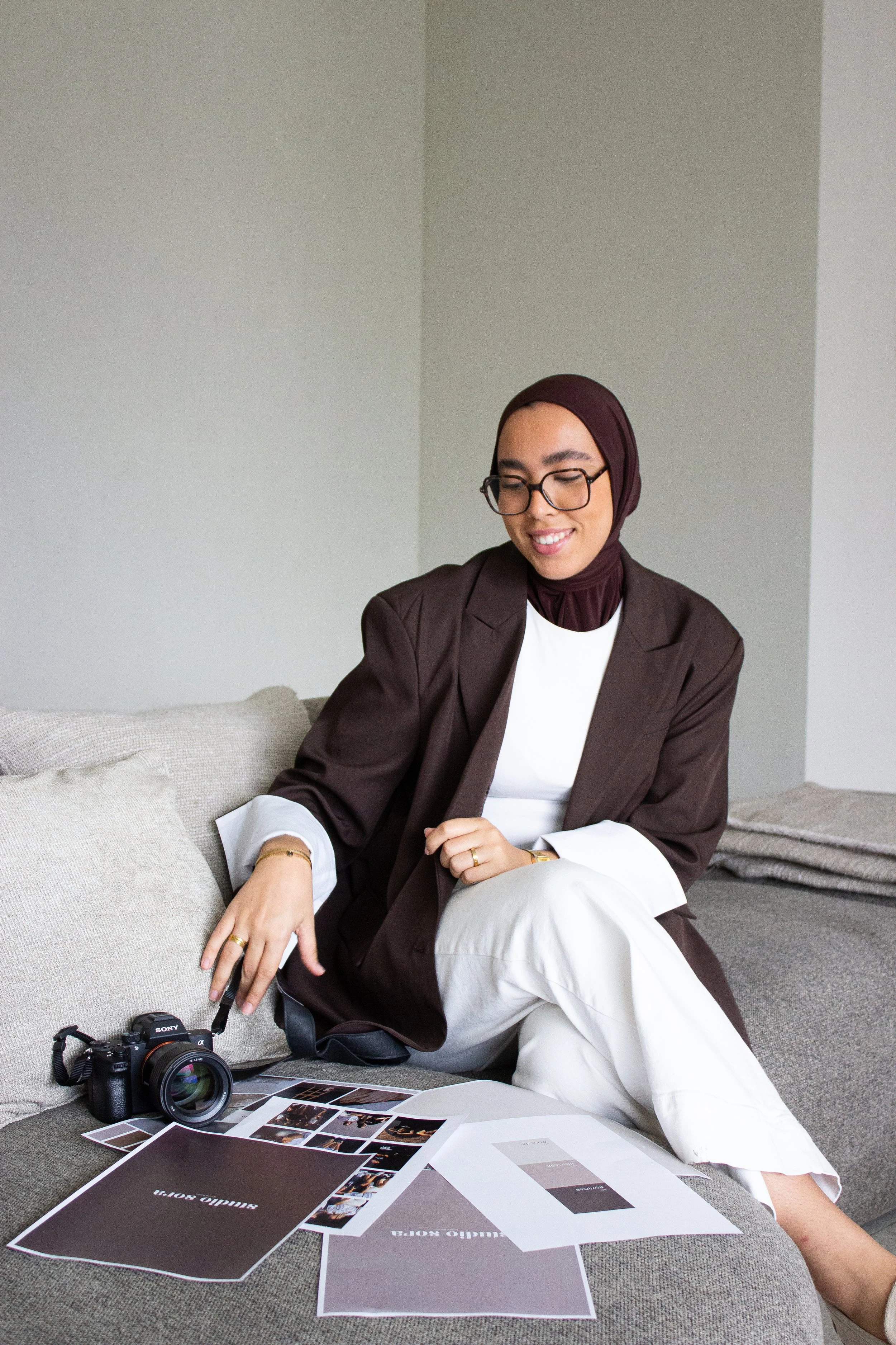 A woman wearing a dark blazer, white pants, and glasses sits on a beige sofa with a camera and printed photos and color samples on her lap.