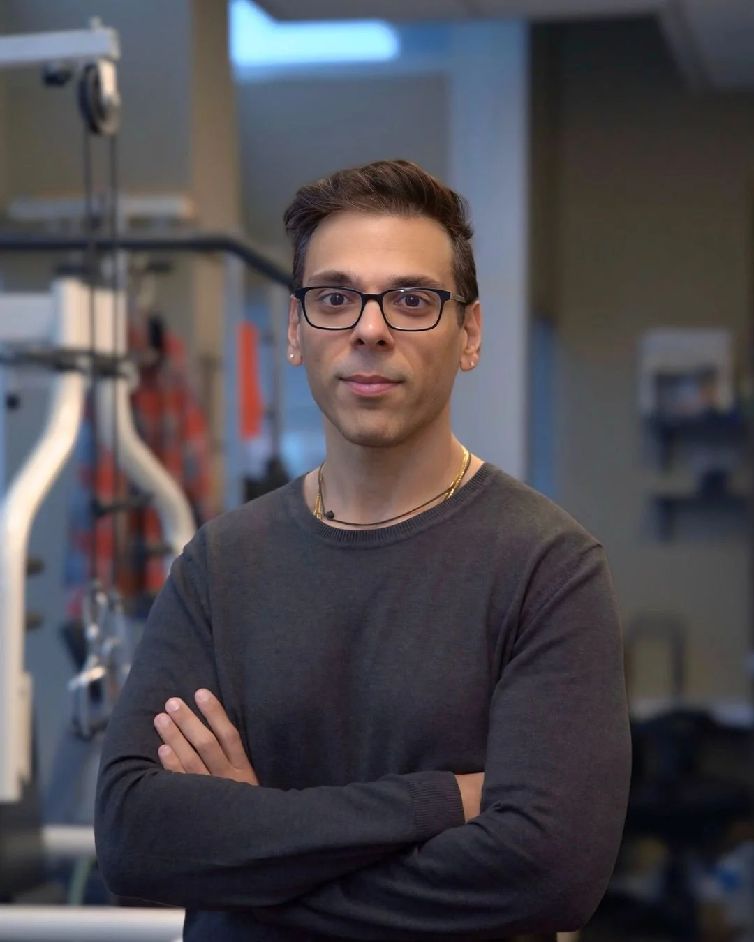 A man with short dark hair, glasses, and earrings standing with arms crossed in a room with medical or laboratory equipment visible in the background.