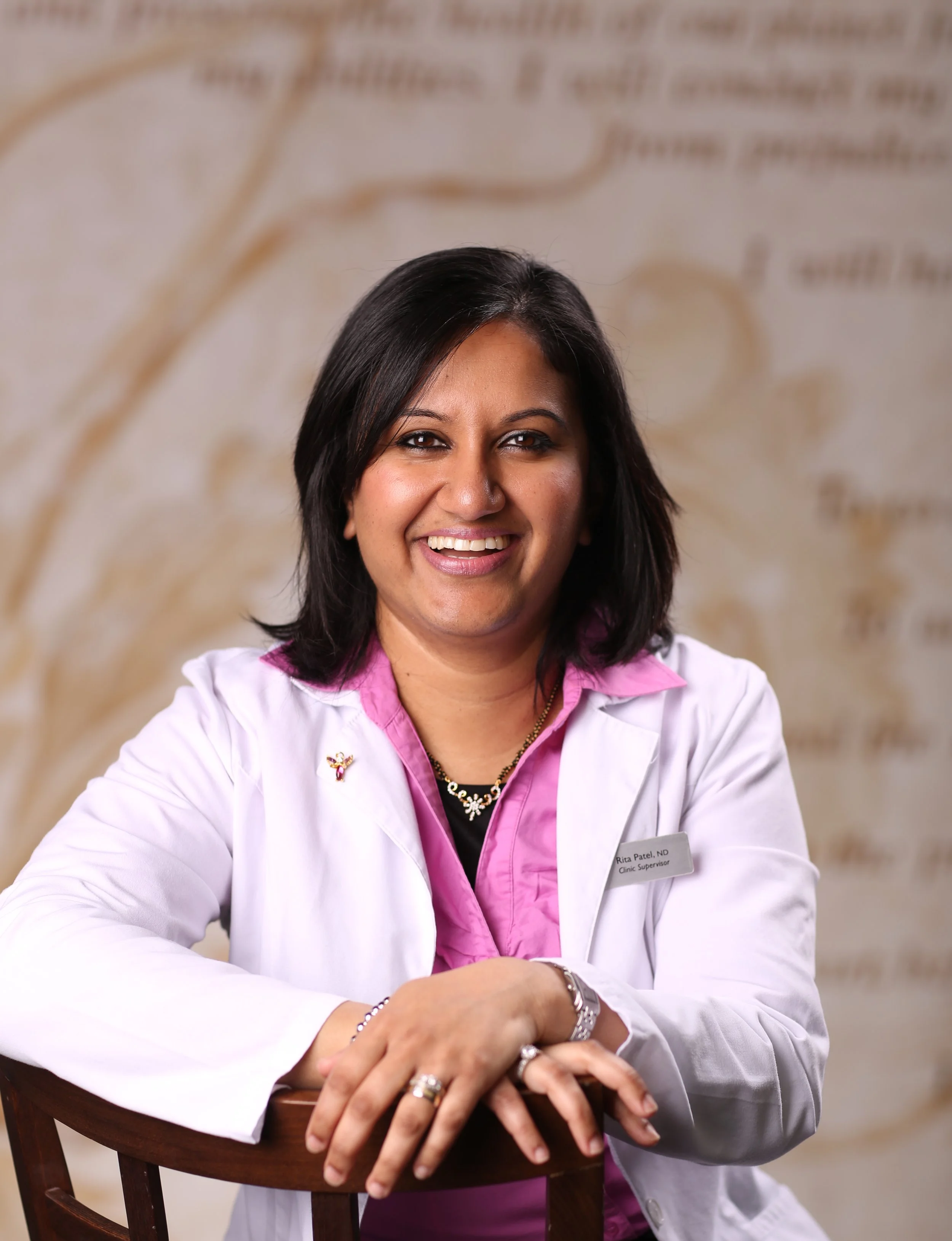 Smiling woman in a white lab coat with pink shirt, sitting at a wooden chair, in front of a blurred background.