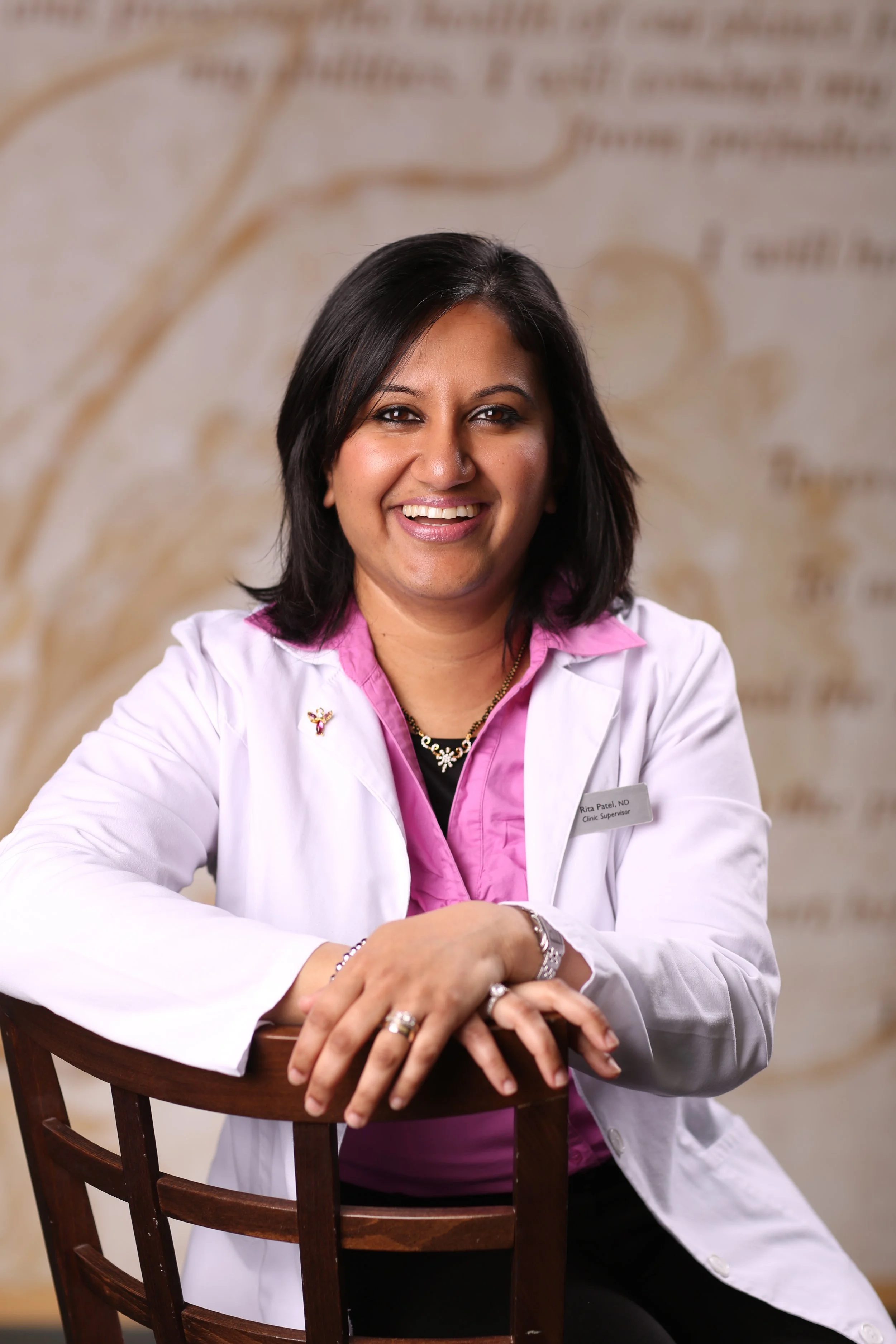 A smiling woman in a white lab coat sitting at a wooden table, with a background featuring text and a faint graphic.