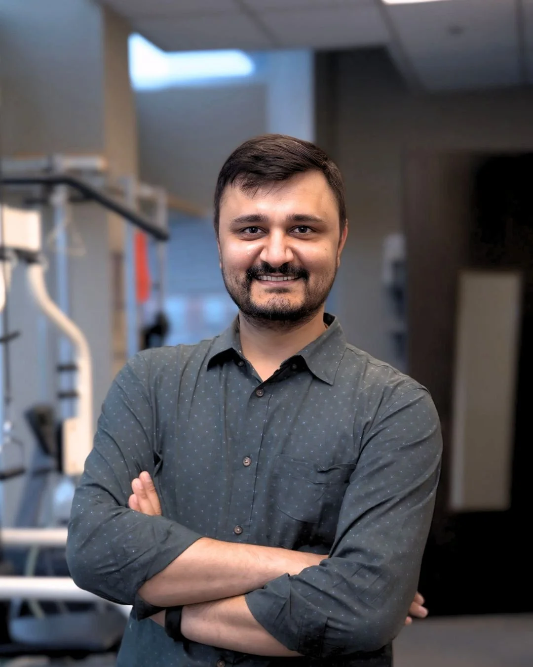 A smiling man with dark hair and a beard, wearing a dark gray button-up shirt, standing with arms crossed in an indoor setting that appears to be a gym or fitness center.