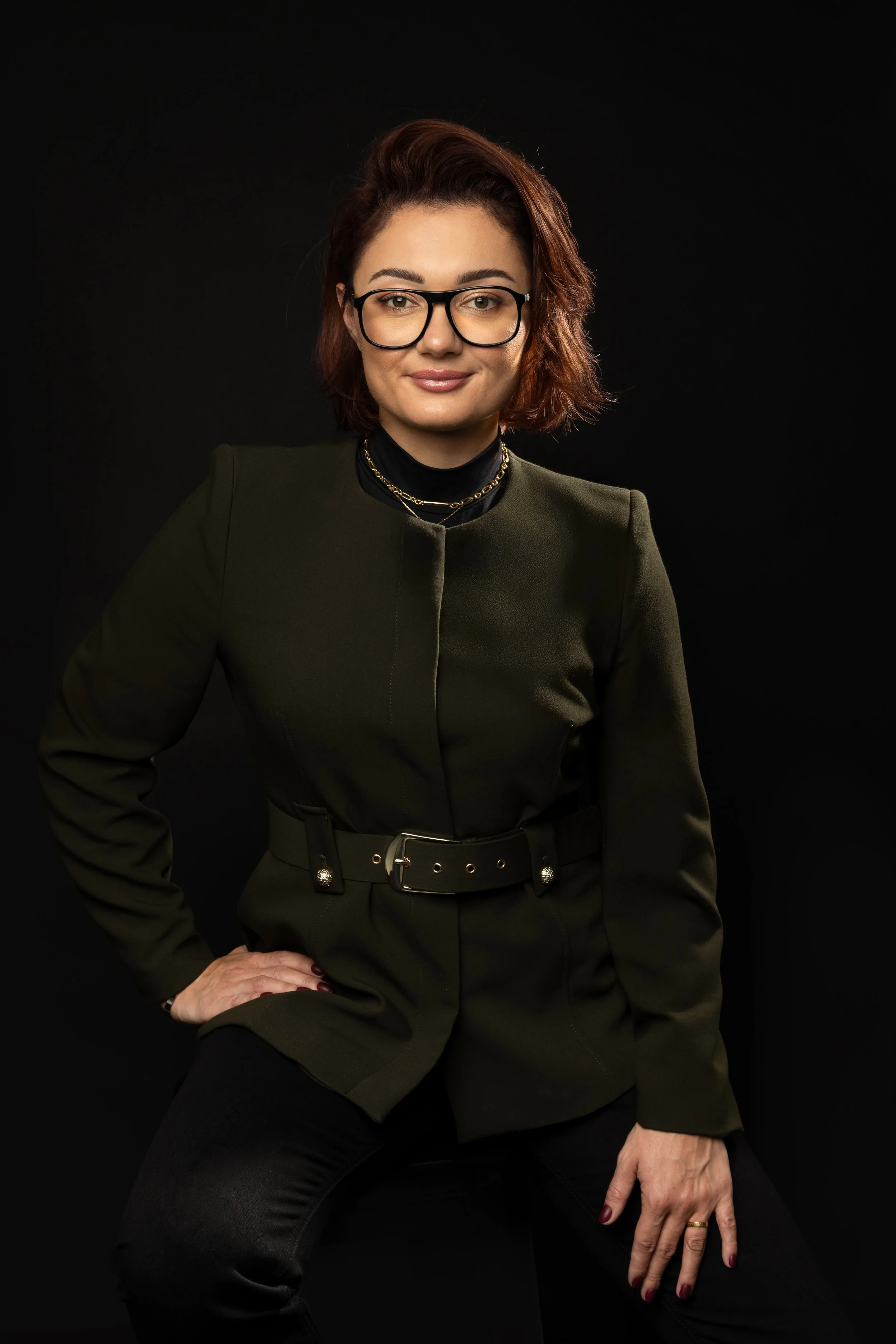 A woman with short auburn hair, wearing glasses and a dark green blazer, sits against a black background. She has a confident expression and is dressed stylishly, accessorized with layered necklaces and a belt.