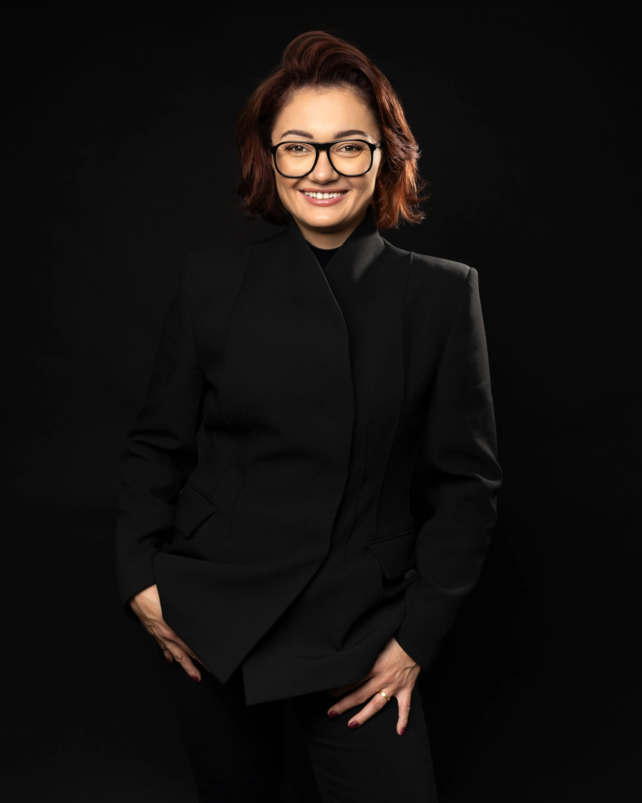 A woman with short red hair, glasses, and a black blazer smiling against a black background.
