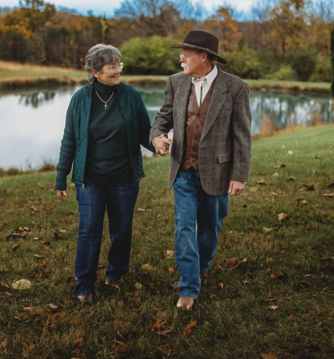 An elderly couple walking hand-in-hand along a grassy area near a lake, with trees showing autumn colors in the background.