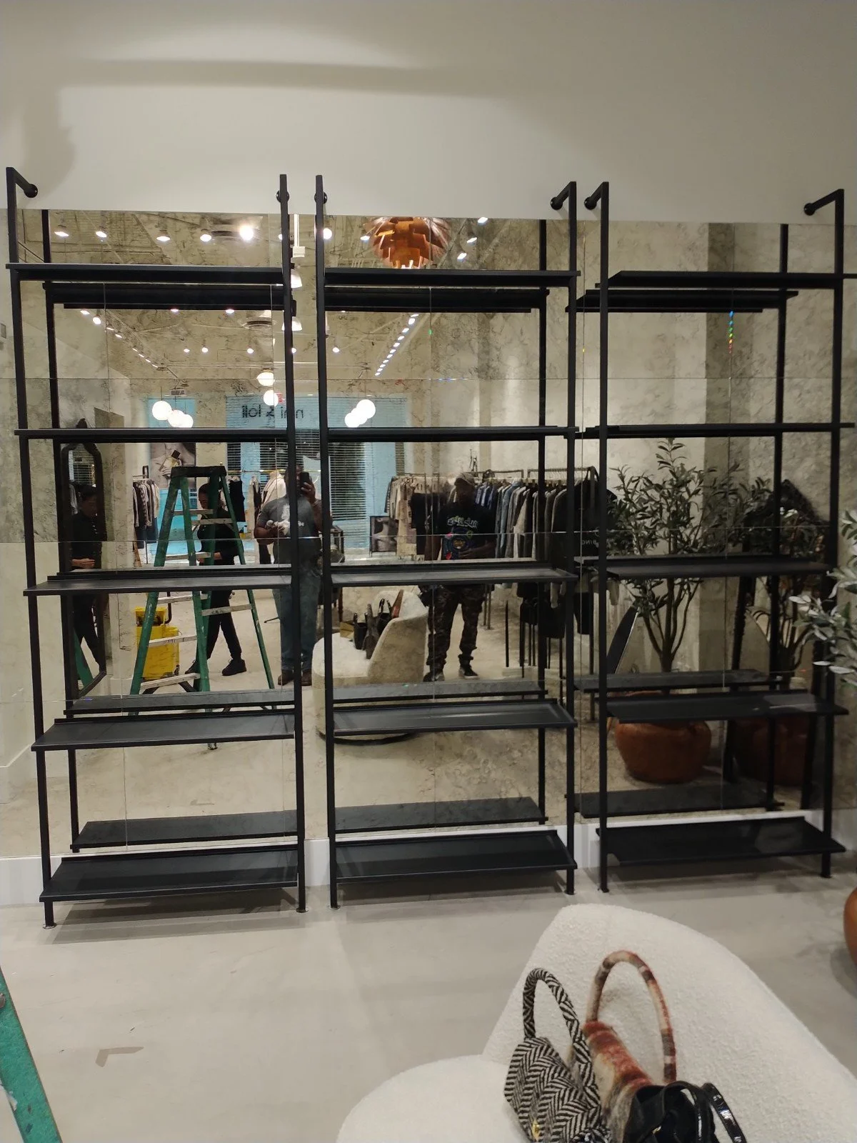 Empty black metal shelving unit in front of a mirror at a clothing store, with clothing racks and shoppers visible in the reflection.