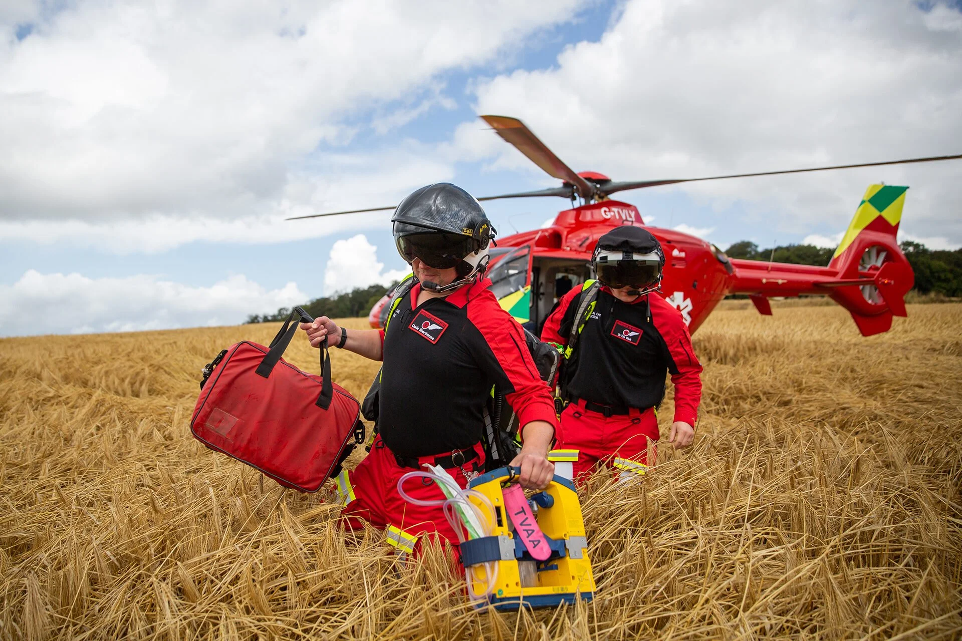 Emergency rescue team members wearing helmets and red uniforms, walking through a wheat field with a red rescue helicopter in the background.