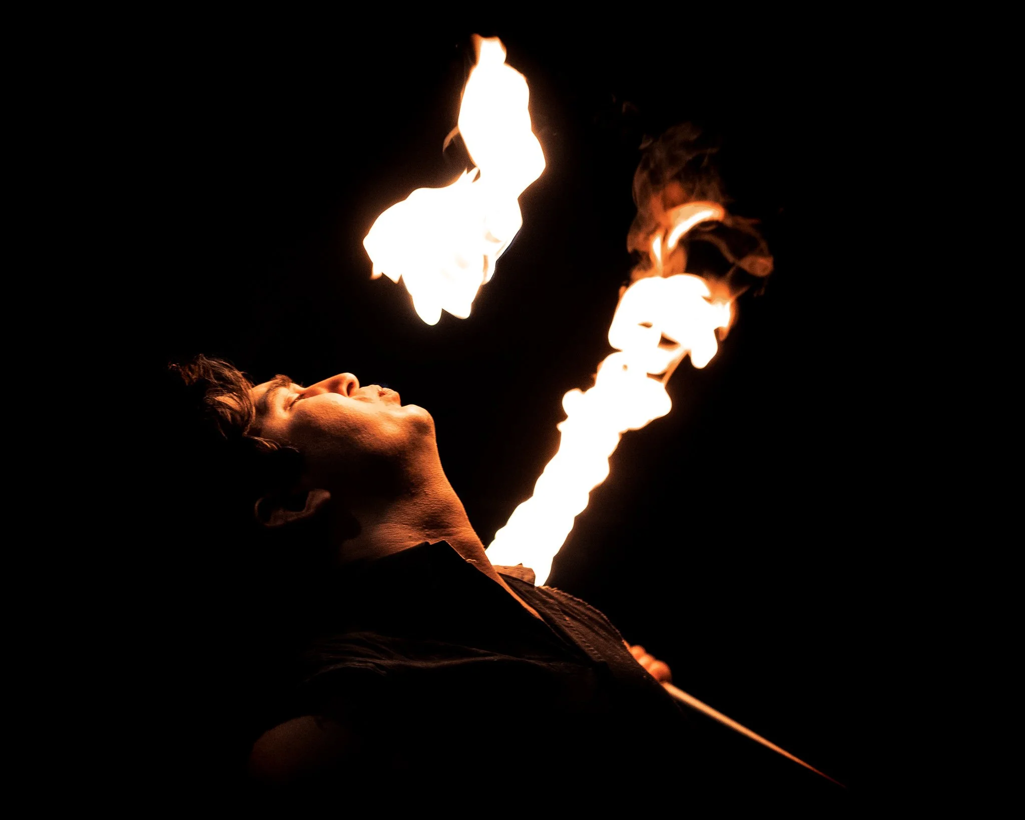 A man performing a fire breathing act at night, with flames erupting from his mouth against a dark background.