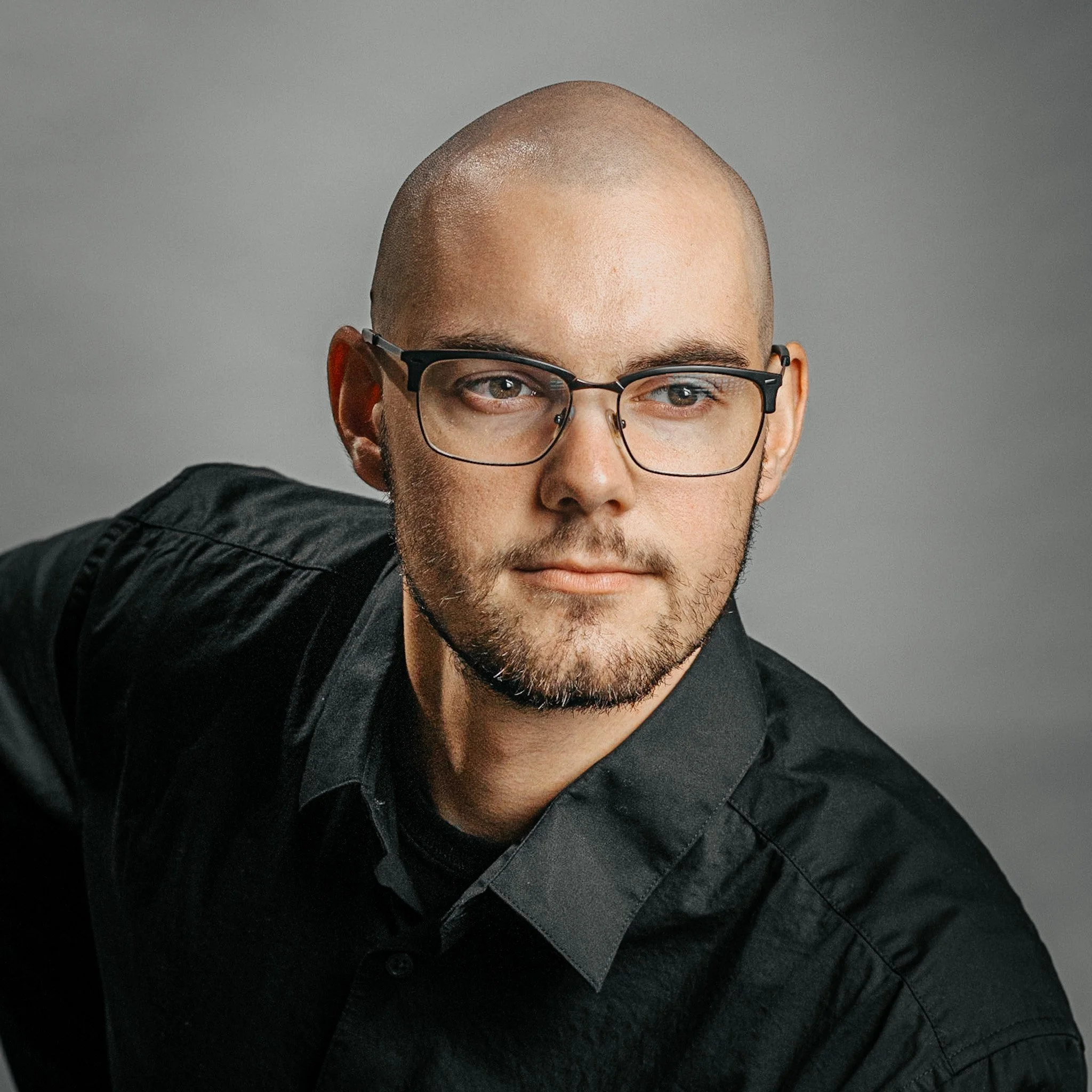Close-up of a man's face, showing the upper part including bald head, glasses, and part of his beard, with a neutral gray background.