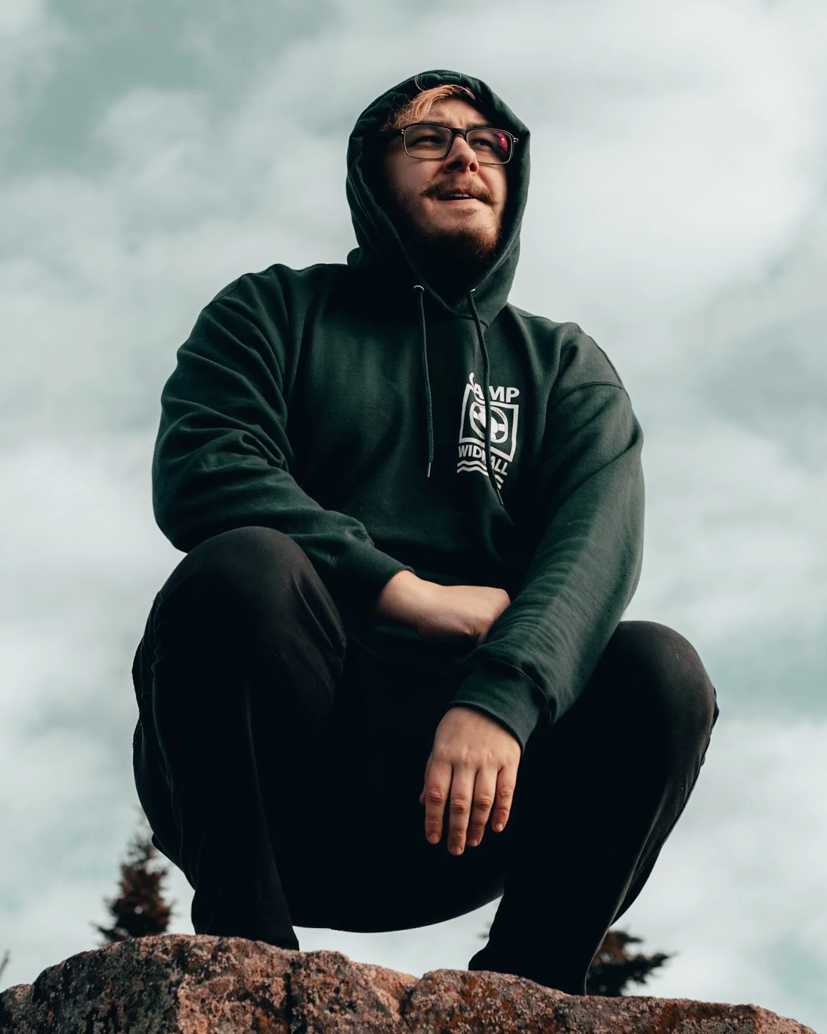 Young man with glasses and facial hair wearing a black hoodie with a logo, crouching on a rock outdoors under cloudy sky.