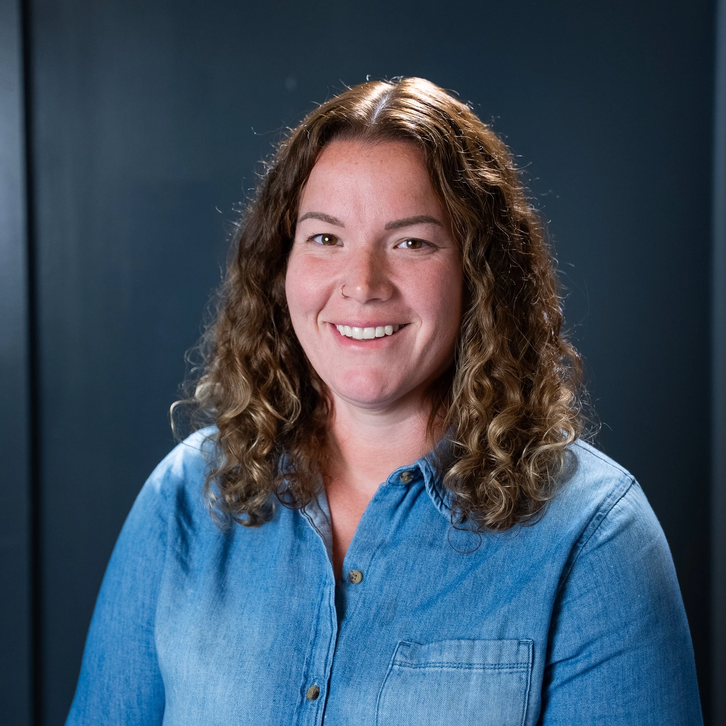 Close-up of a woman with curly brown hair, fair skin, wearing a blue denim shirt, smiling, against a dark background.