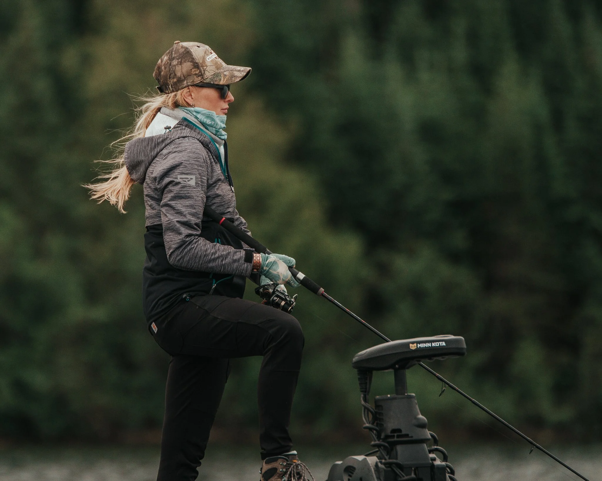 A woman riding an electric bicycle outdoors in a green, wooded area. She is wearing a camouflage hat, sunglasses, a gray jacket, and a neck gaiter, with her blonde hair tied back.