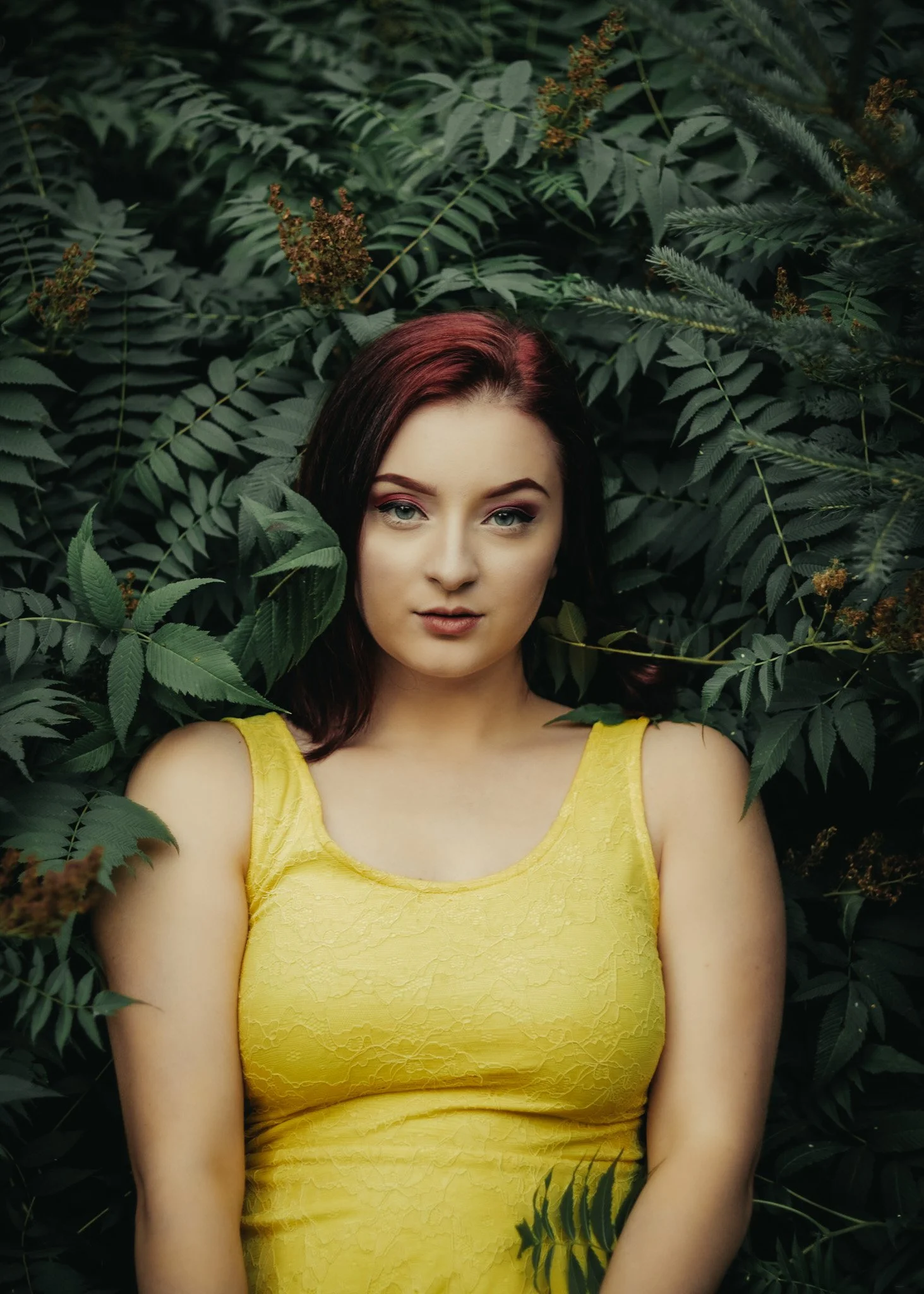 A woman with dark red hair wearing a yellow dress, surrounded by green leafy plants.