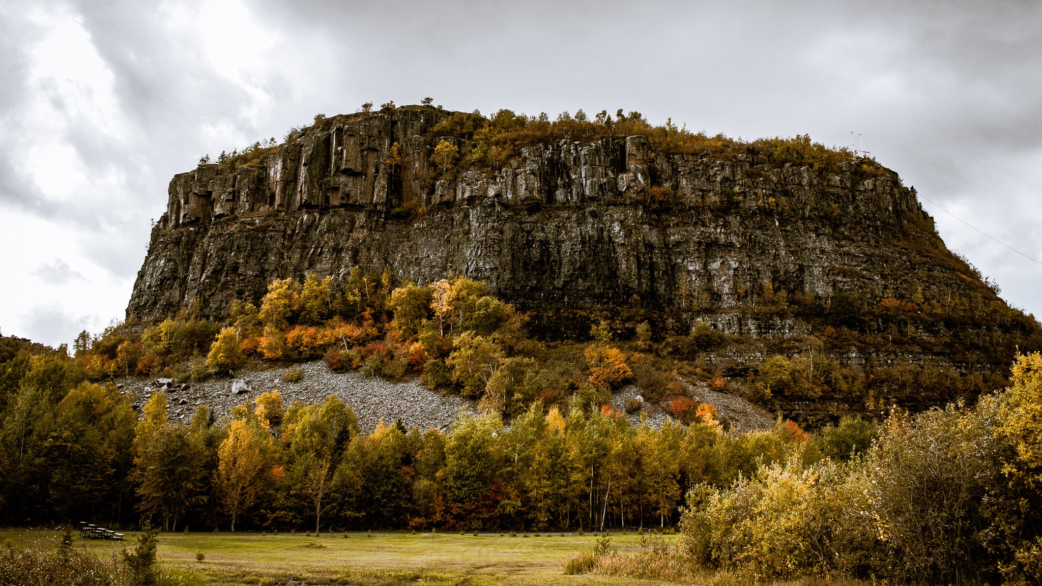 Large rocky hill with steep rock faces, surrounded by autumn-colored trees and a grassy field in the foreground, with a cloudy sky overhead.