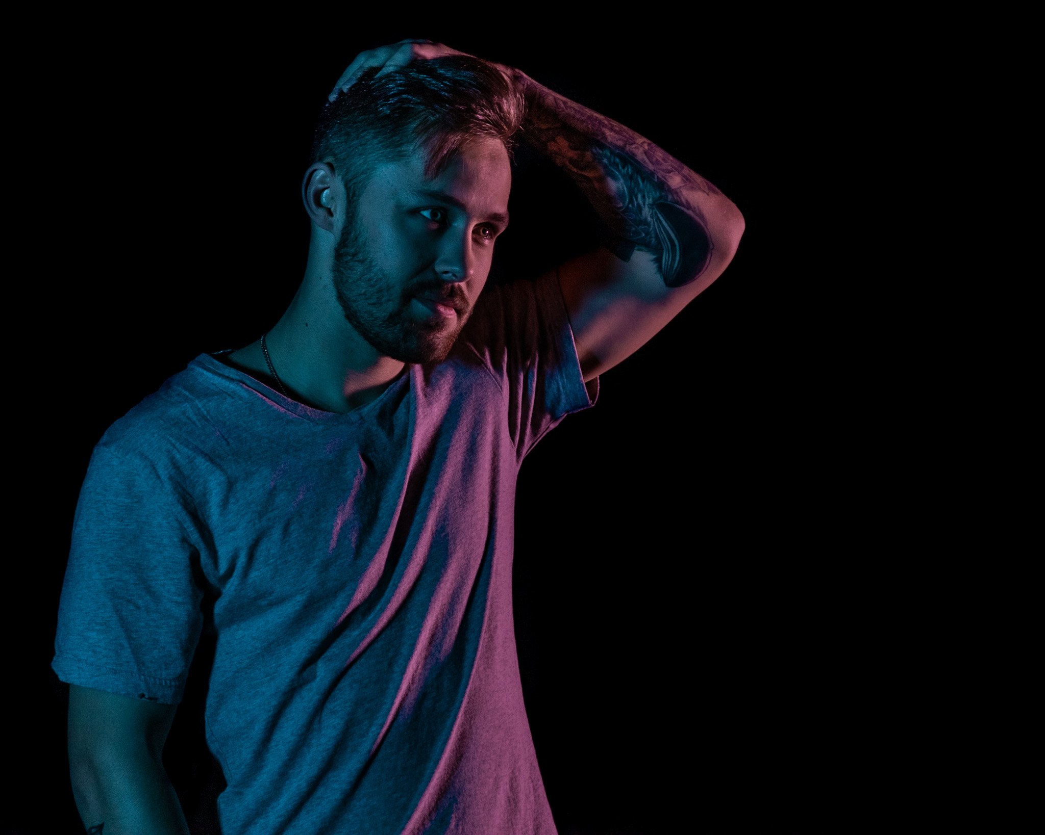 A young man with tattoos on his arm, wearing a gray T-shirt, is holding his hair back with his hand under a dark background with colorful lighting.