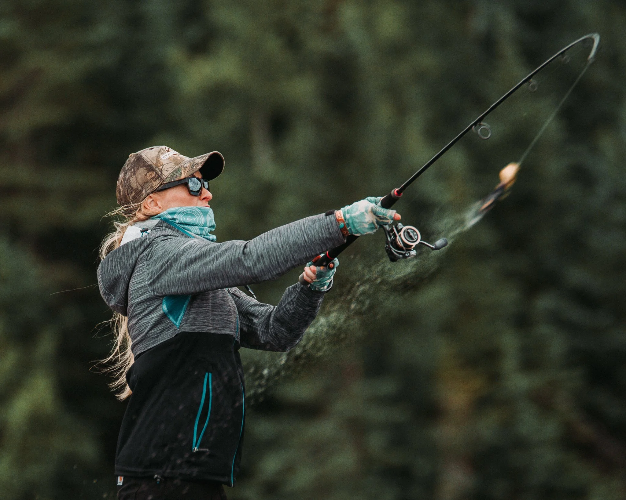 A woman fishing outdoors, wearing sunglasses, a camouflage hat, a gray jacket, and gloves, holding a fishing rod with water droplets on it.